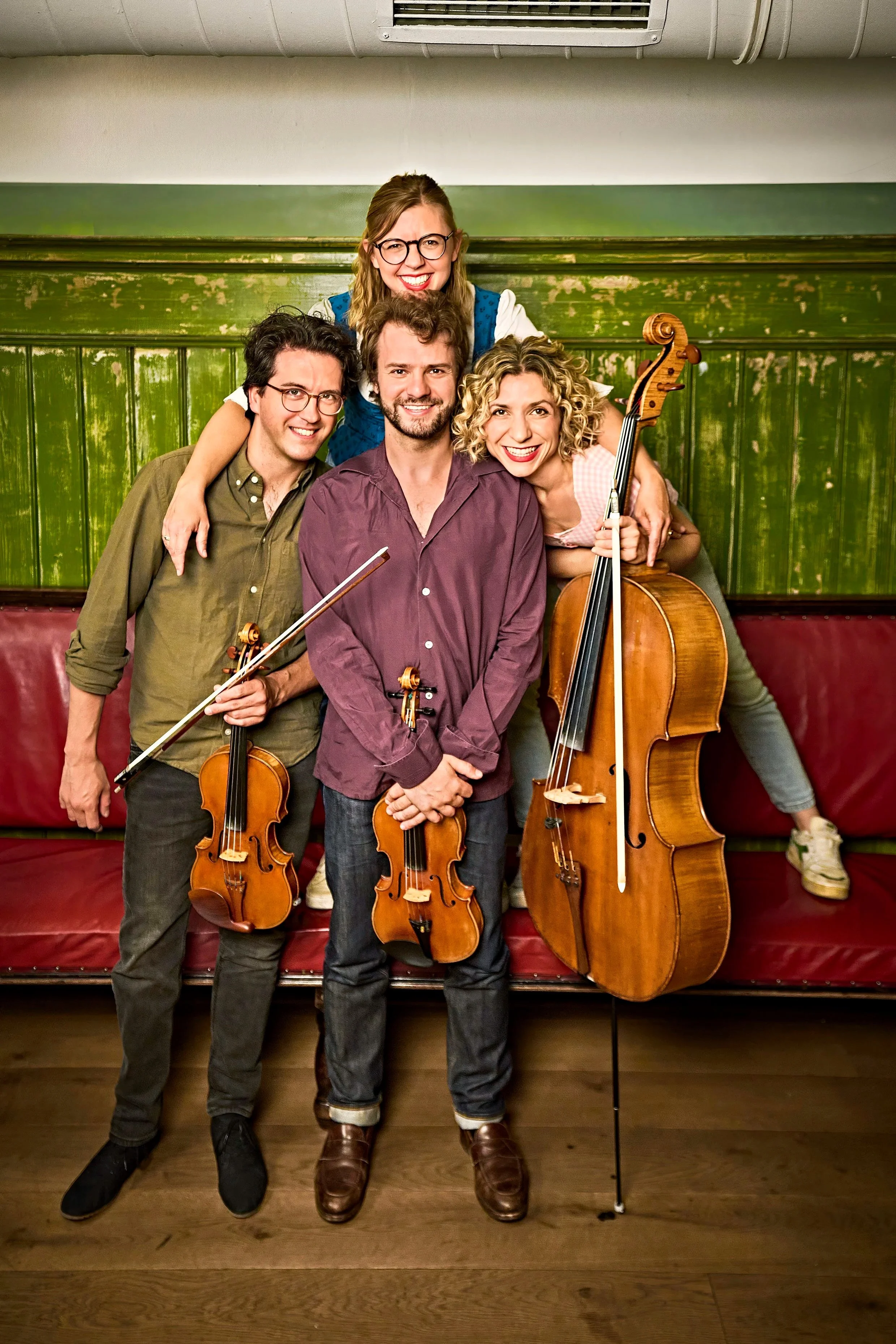 Four smiling musicians pose with two violins and a cello in a vintage green-paneled room.
