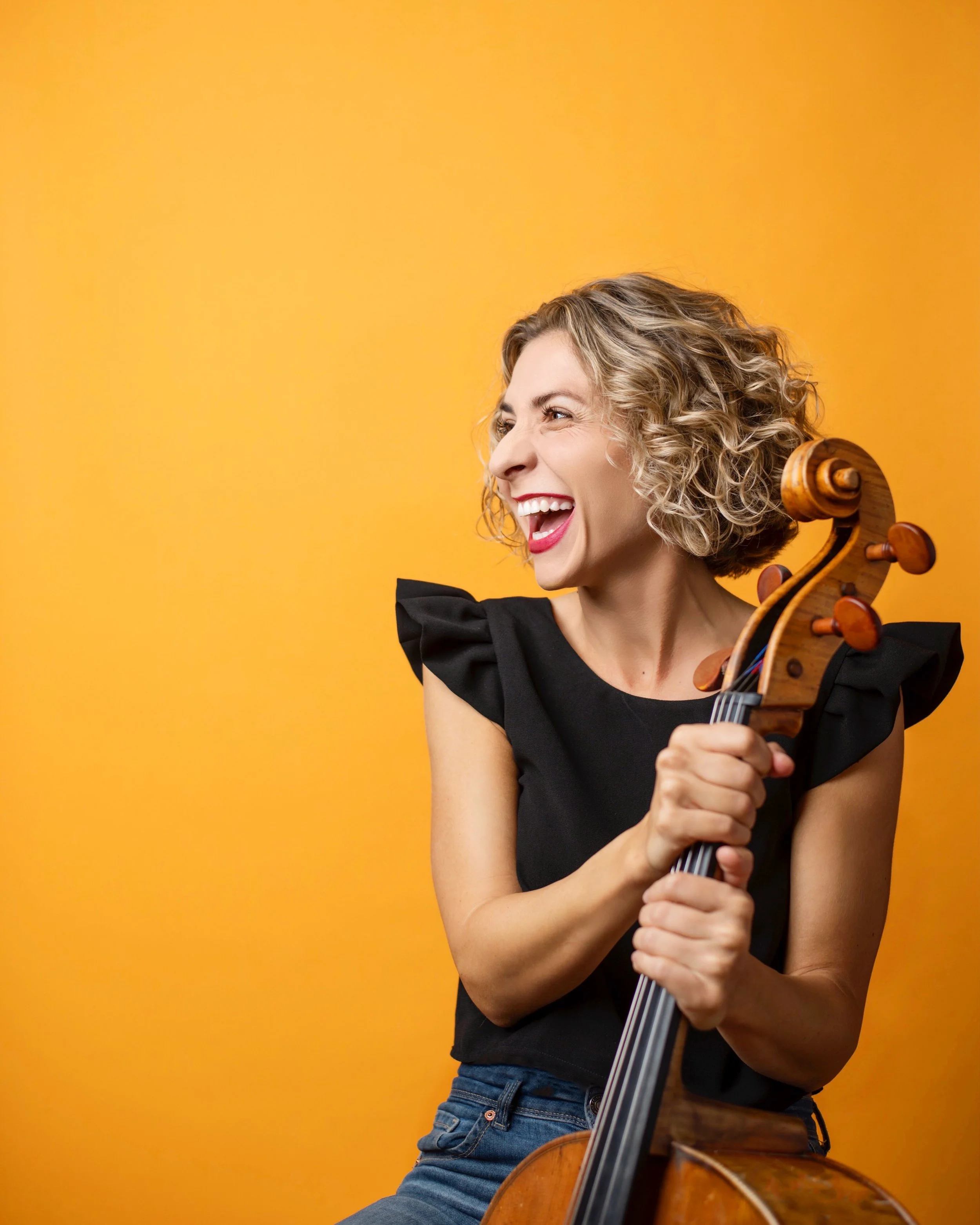 Smiling woman with curly hair holding a cello against a bright orange background