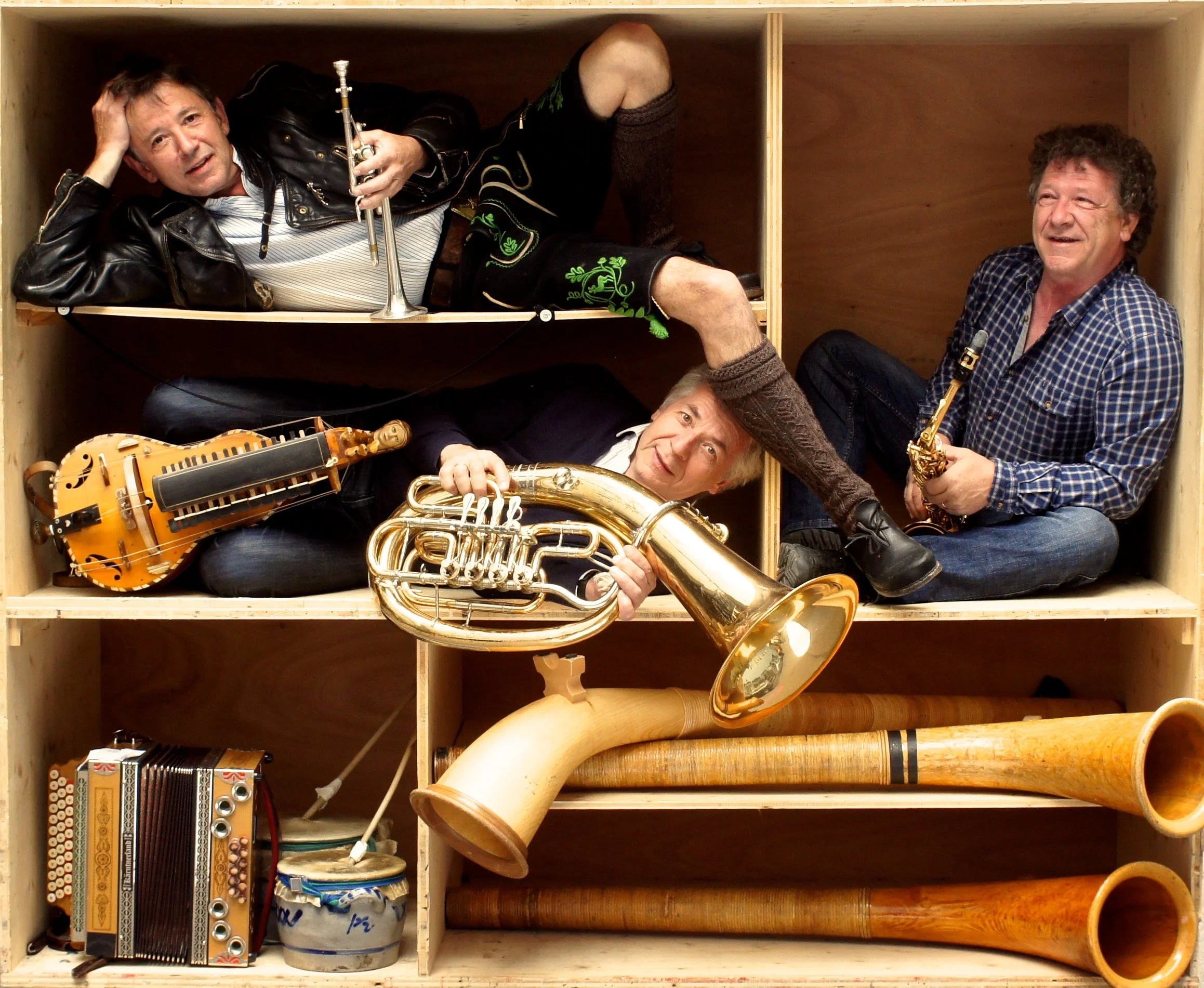 Four musicians pose inside wooden shelving cubbies with brass instruments, a hurdy-gurdy, accordion, drum, and alpine horns.