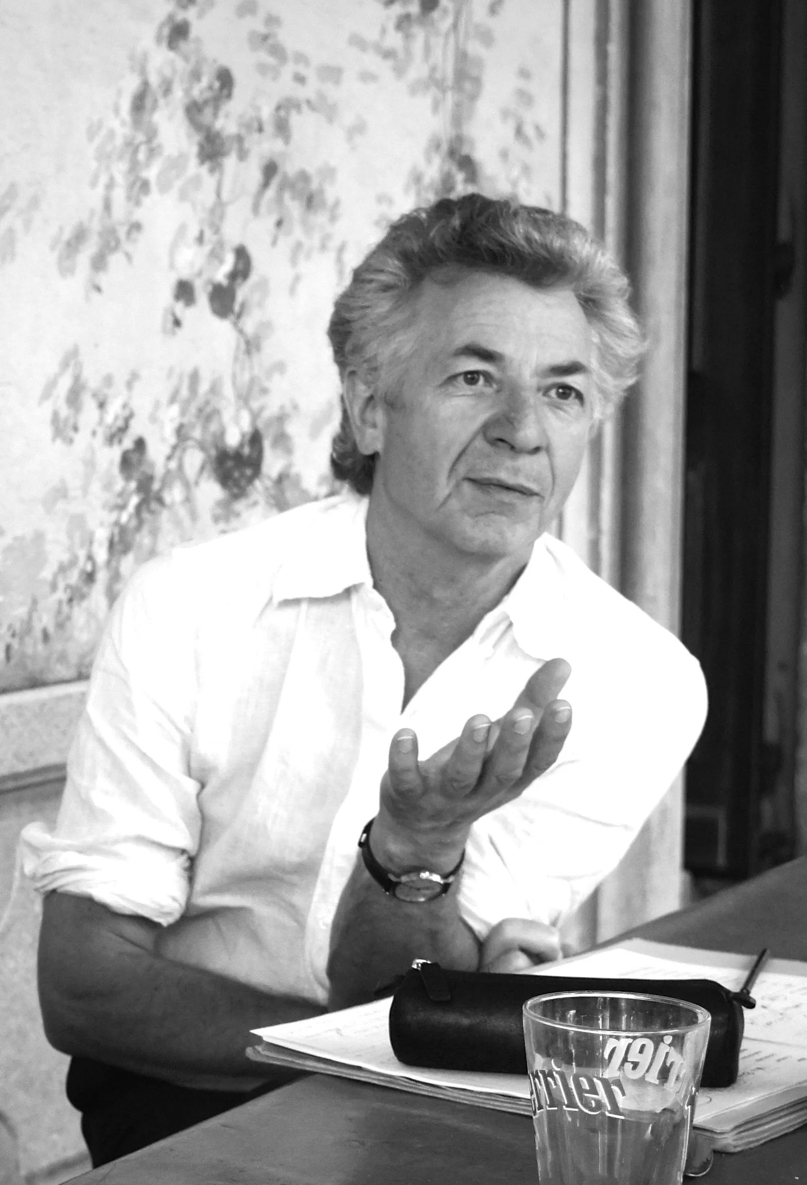 Black-and-white portrait of an older man in a white shirt gesturing at a table with papers, a case, and a drinking glass.