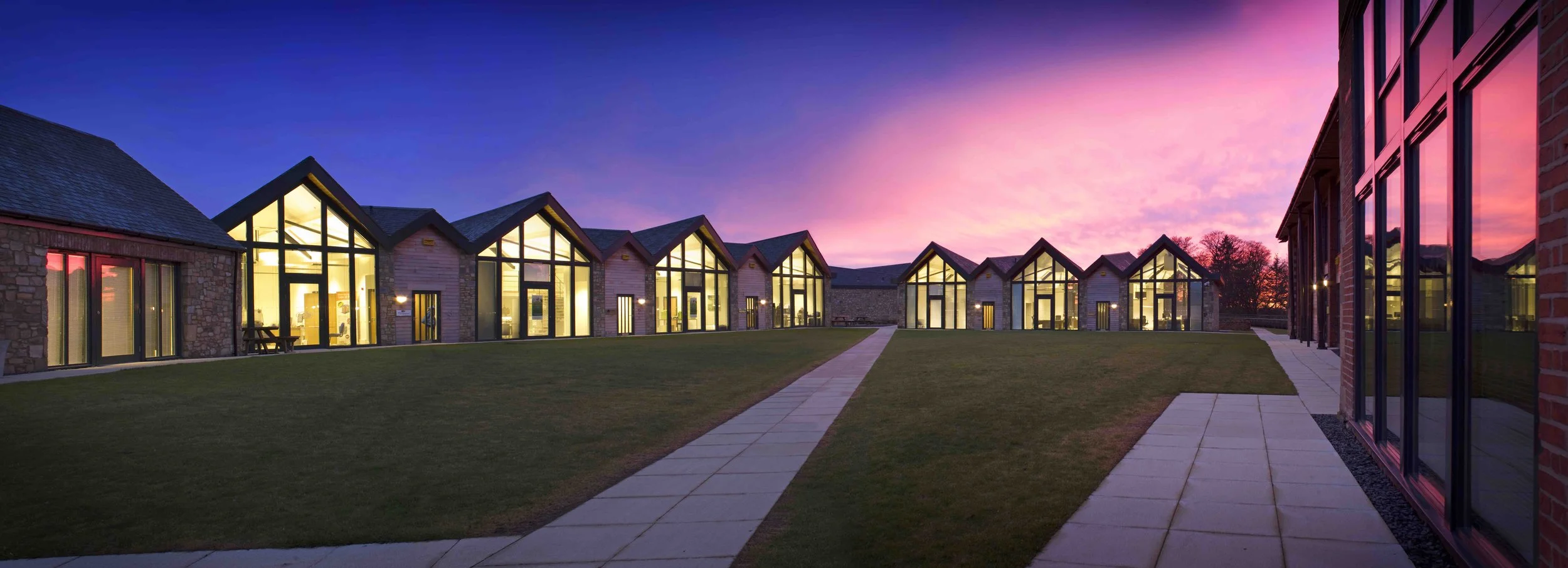 Modern office buildings at Prestwick Parkwith large glass windows and peaked roofs during sunset, with a grassy yard and concrete pathway in the foreground.