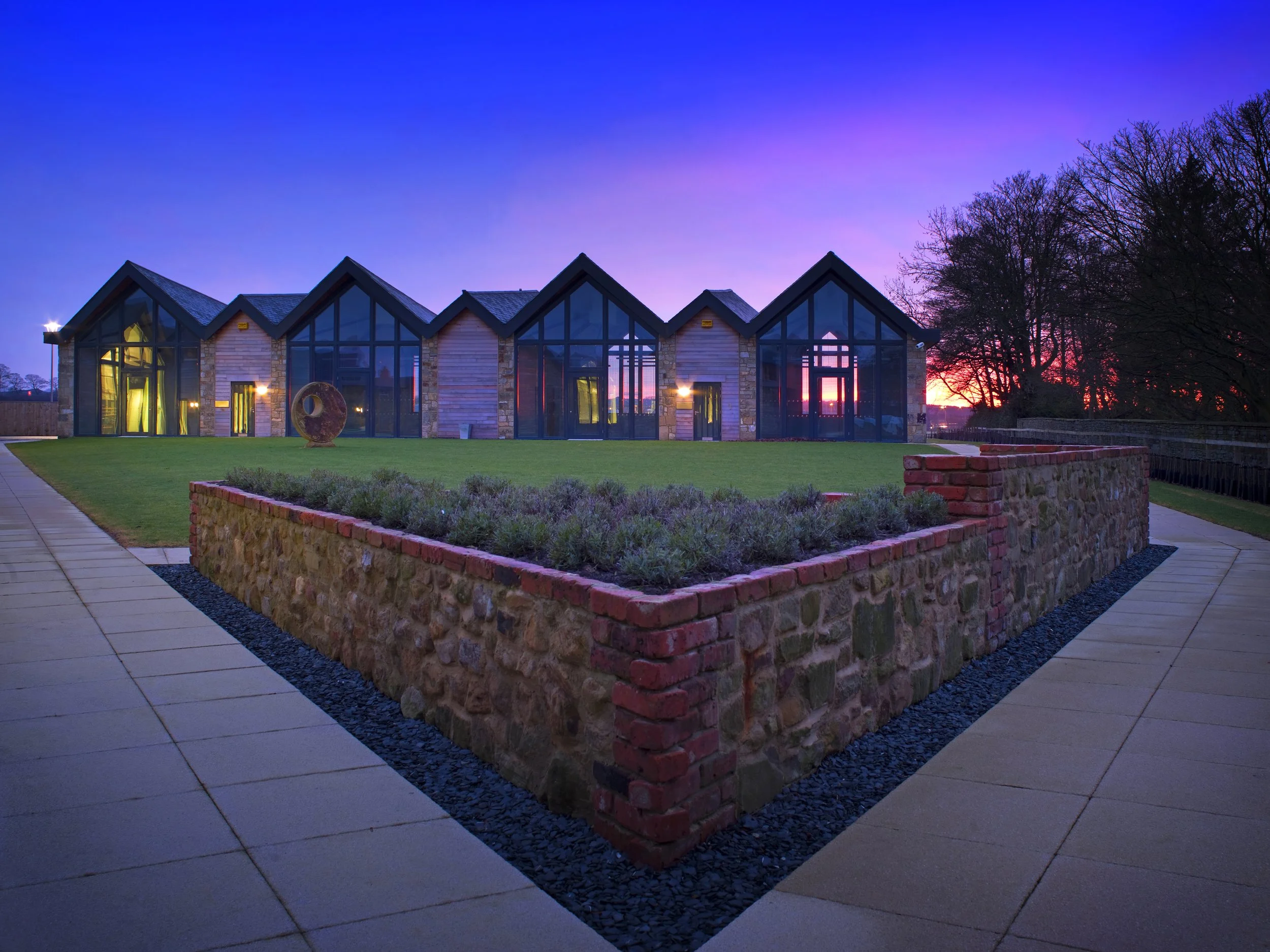 A modern office building at Prestwick Park with large glass windows and a brick exterior under a colorful sunset sky, surrounded by a paved walkway and a low brick wall.