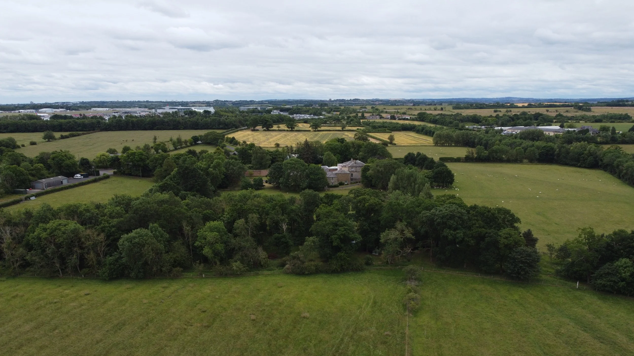 A scenic aerial view of lush green fields, trees, and scattered houses under a cloudy sky.