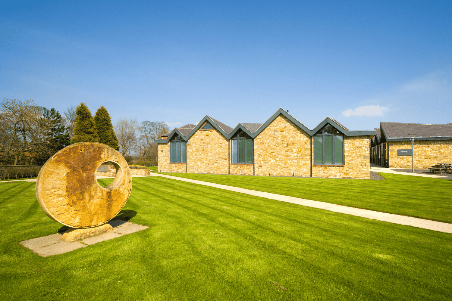 Modern office building with stone walls and large windows, situated on a well-maintained grassy area with a concrete pathway.
