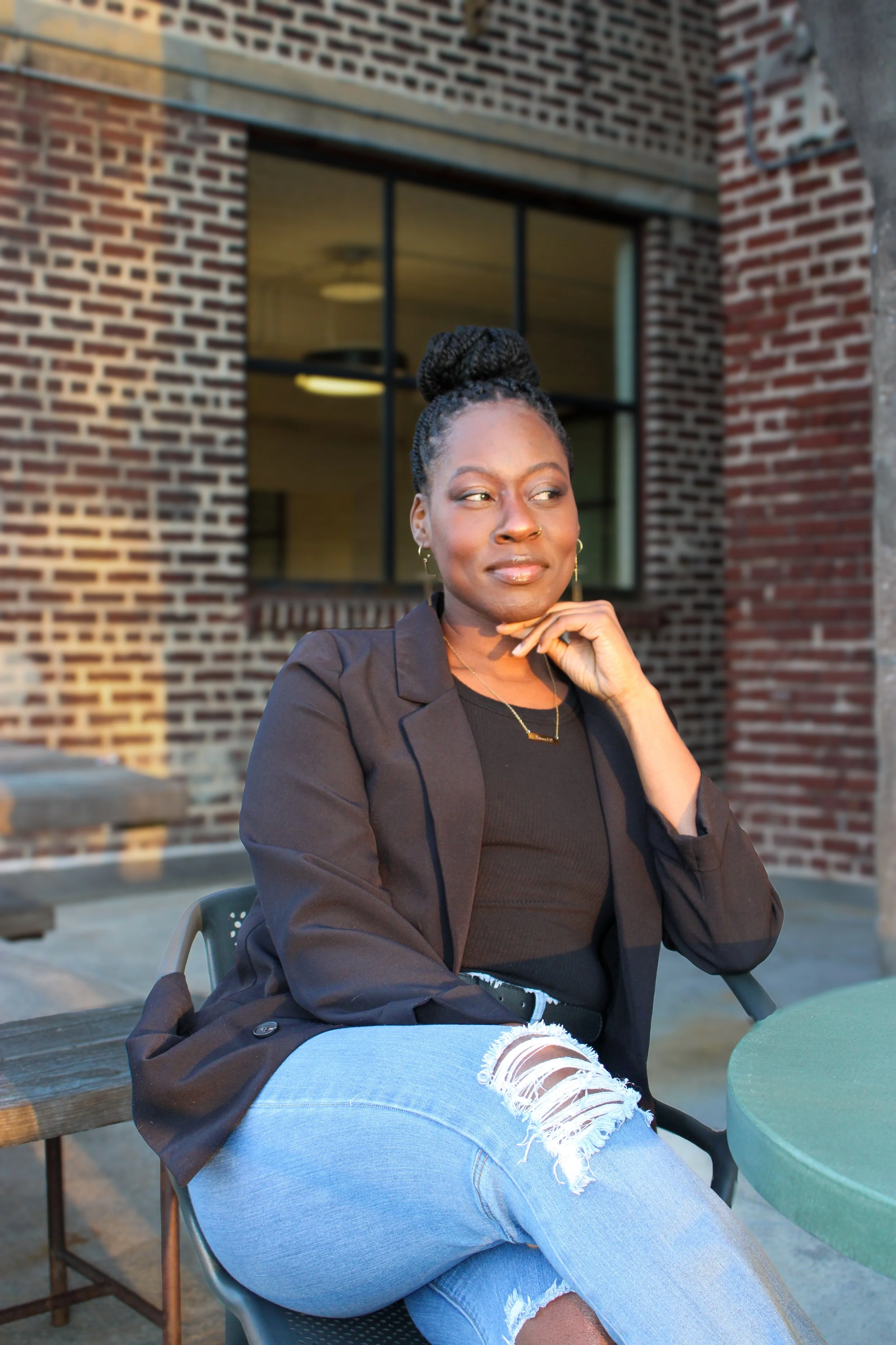 A black woman sitting outdoors at a brick building, wearing a black blazer, black top, and distressed light blue jeans, with her hair in an updo and her hand resting on her chin, looking thoughtful.