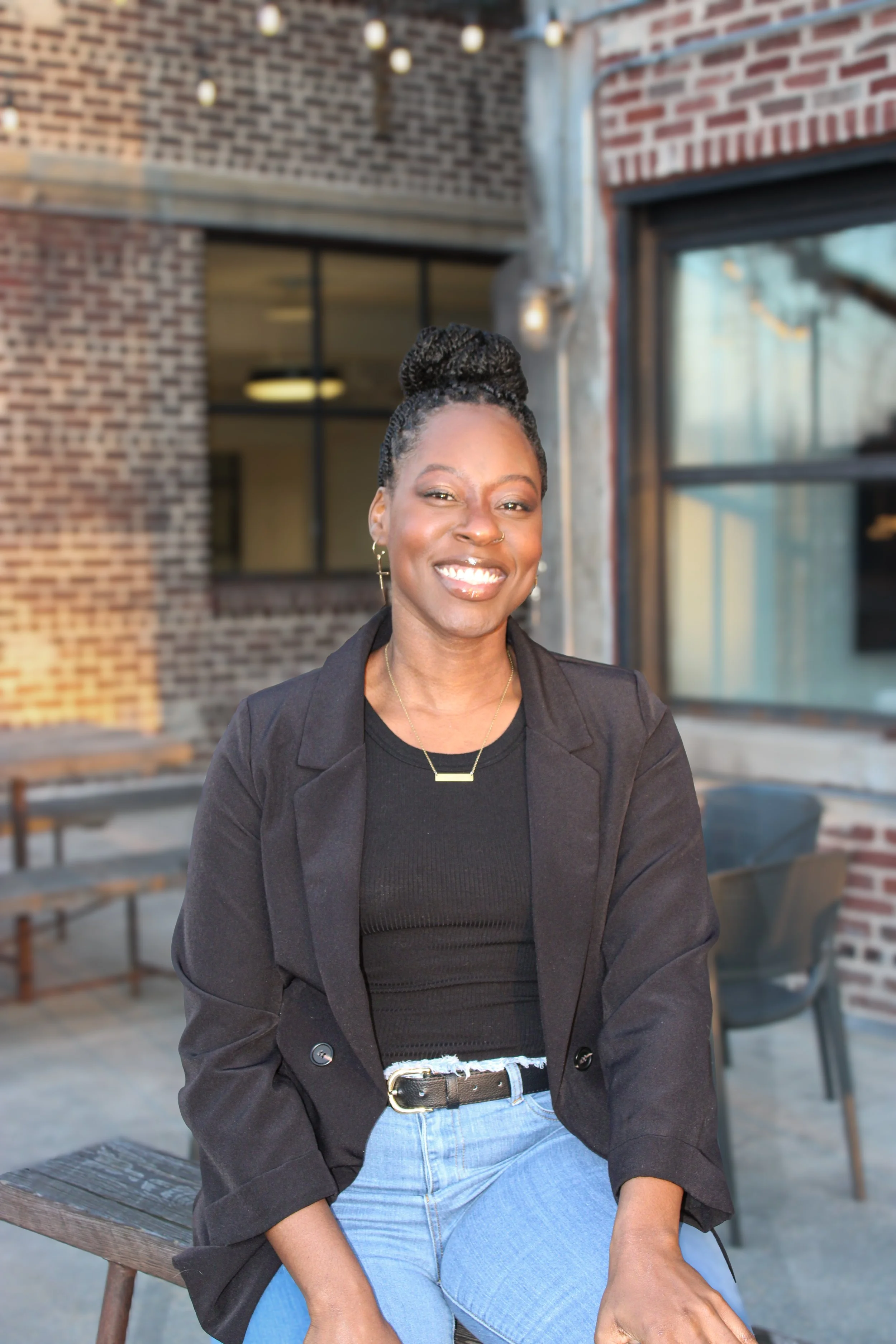 A smiling Black woman with her hair styled in a bun, sitting outdoors at a restaurant or café with brick walls and string lights overhead.