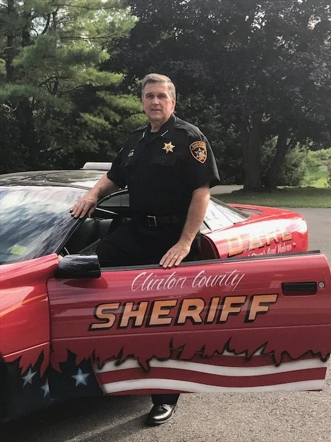 Sheriff Dave Favro in uniform, standing with one hand on the open door of a red Clinton County sheriff's patrol car.