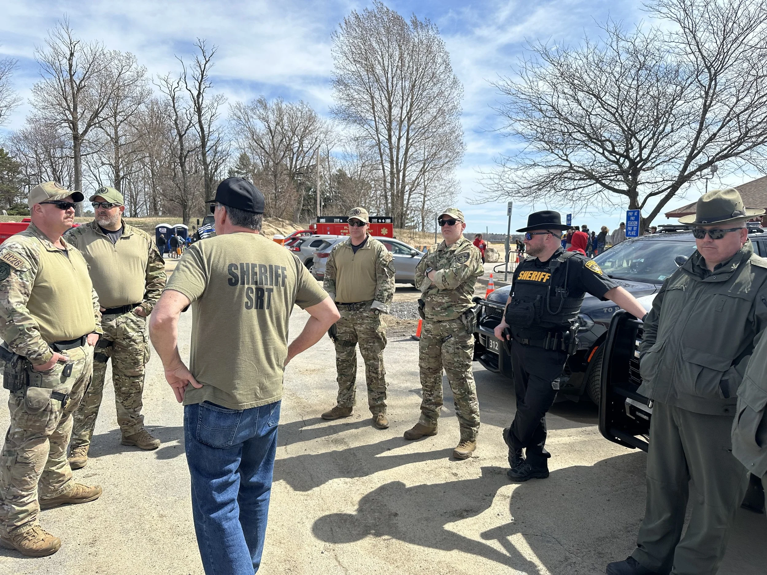 Group of law enforcement officers and emergency personnel standing and talking outdoors near parked vehicles on a clear day with leafless trees in the background.