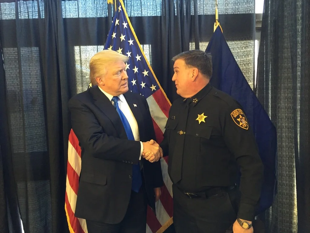 President Donald Trump shaking hands with Sheriff Dave Favro in front of American flags.