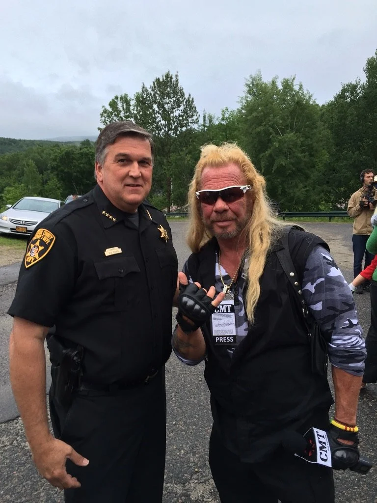 Sheriff Dave Favro and Dog the Bounty Hunter are standing outdoors on a cloudy day, with trees and a few people in the background.