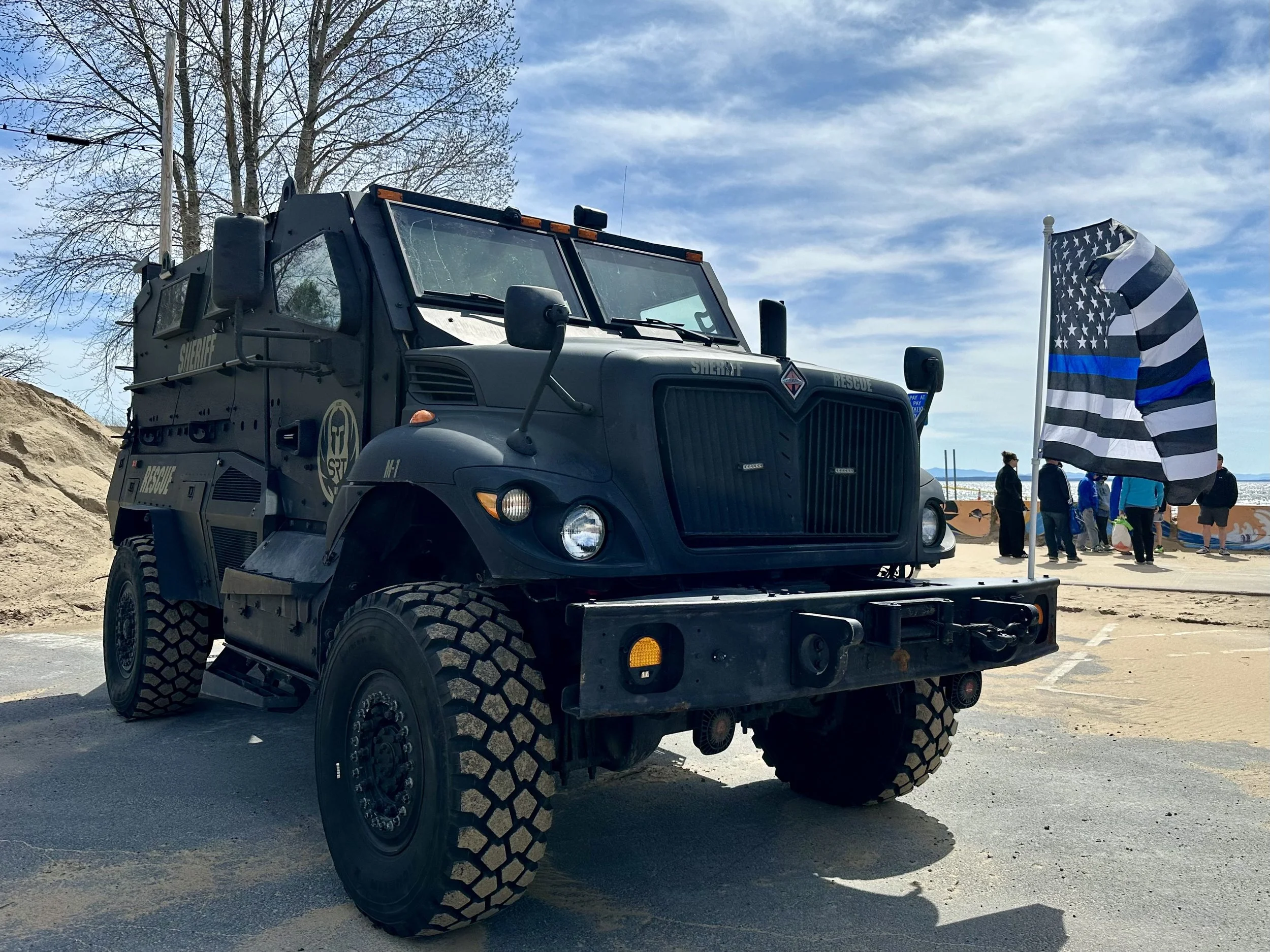 Black Clinton County sheriff rescue vehicle parked outdoors with a blue and black flag on a pole nearby. People are gathered in the background near the ocean under a partly cloudy sky.