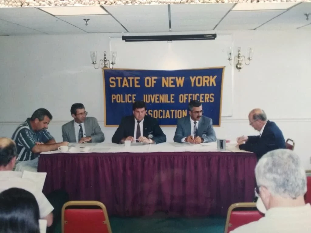A panel of five men sitting at a long table in a conference room during a meeting, with a blue banner behind them that reads, 'STATE OF NEW YORK POLICE JUVENILE OFFICERS ASSOCIATION'.