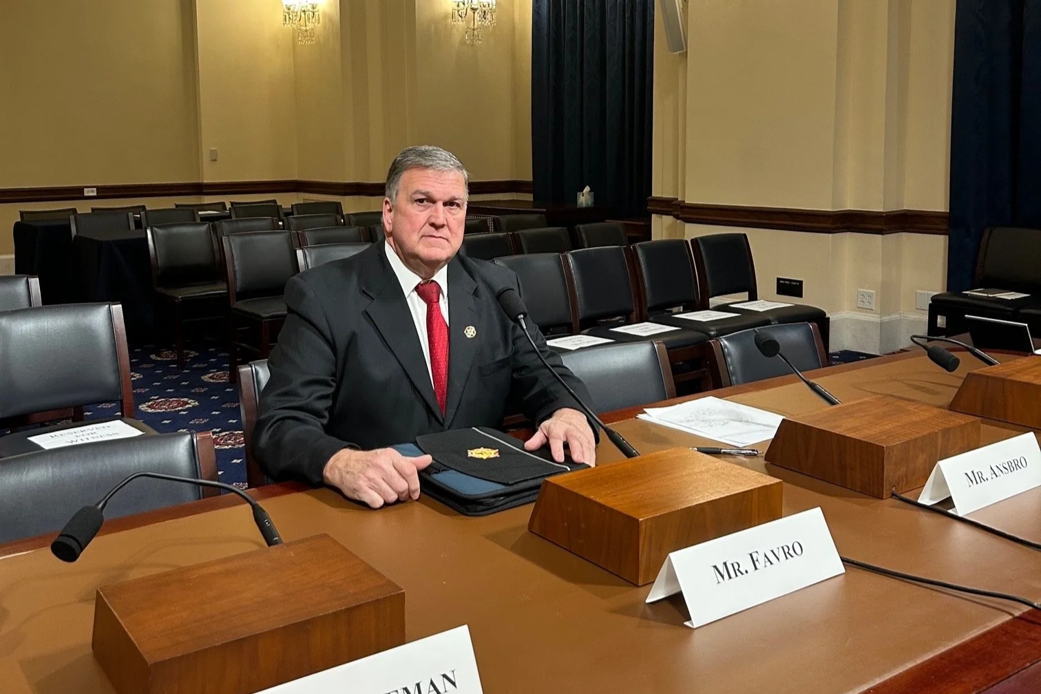 A man in a dark suit, white shirt, and red tie seated at a conference table with a black folder and papers in front of him. The room has empty chairs arranged behind him and nameplates on the table reading "Mr. Favro" and "Mr. Asbro." The room appears to be a formal meeting or hearing room with chandeliers and dark curtains.