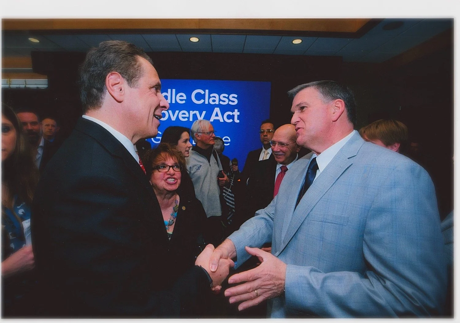 Sheriff Dave Favro and Governor Andrew Cuomo shaking hands and talking at a political event, with a group of people in the background and a blue screen displaying text about the Affordable Care Act.
