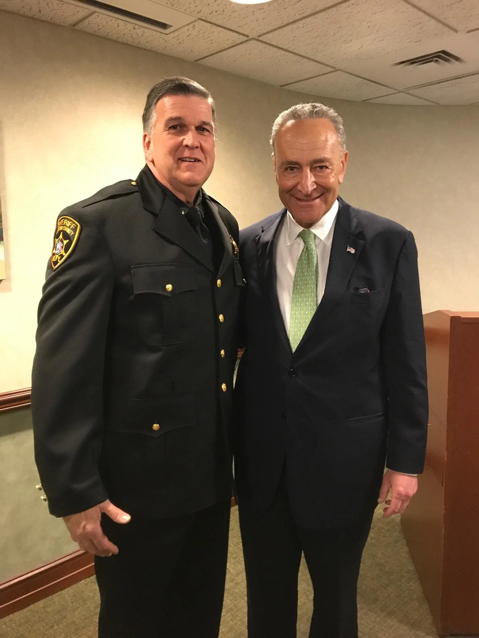 Two men in formal attire standing indoors, one in a sheriff's uniform and the other in a suit with a green tie, smiling at the camera.