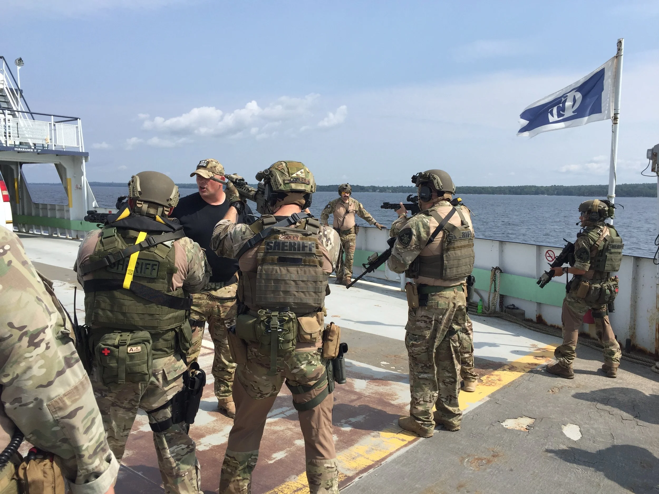 Clinton County Sheriff's Department SRT in camouflage uniforms on a boat, possibly on a water rescue or patrol mission, with Water and a shoreline visible in the background, and a flag flying on the boat.