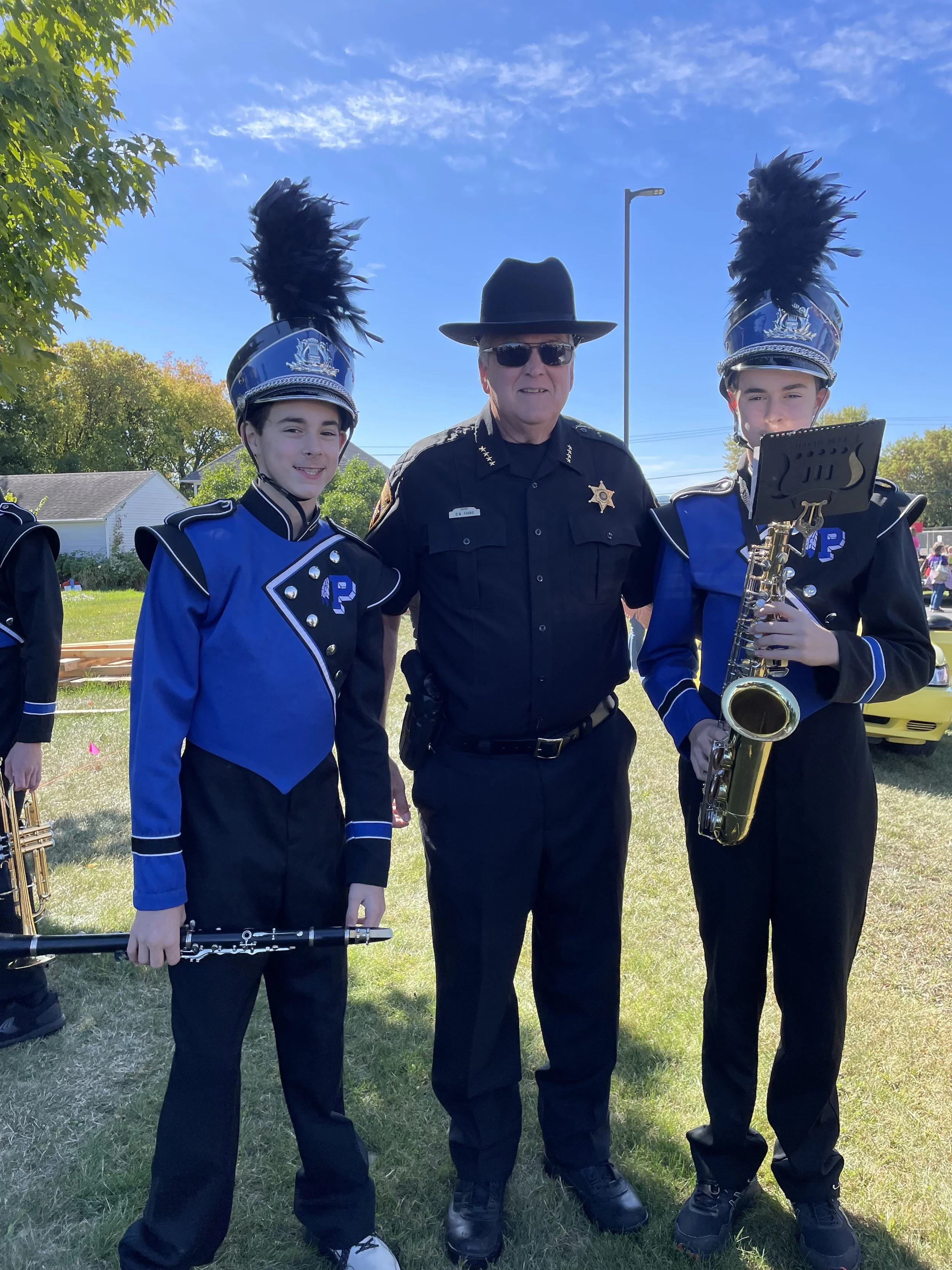 Two young band members in blue marching band uniforms with black feathered plumes on their hats, standing next to a police officer in uniform, outdoors on a sunny day.