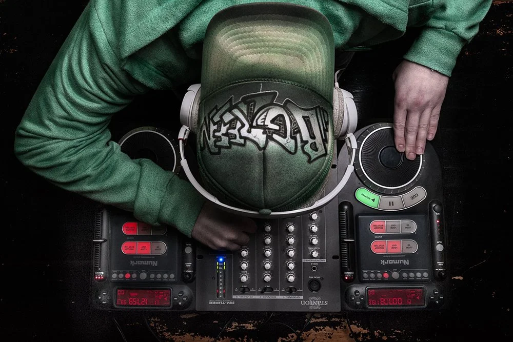 A person wearing a green hoodie and cap with graffiti, using DJ equipment on a black surface.