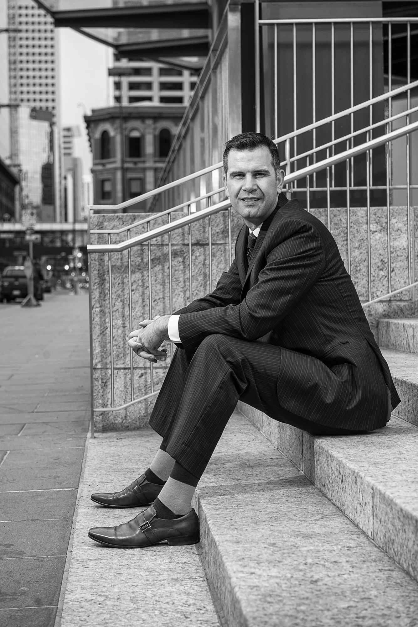A man in a business suit sitting on outdoor steps in an urban setting, smiling at the camera, with tall buildings in the background.