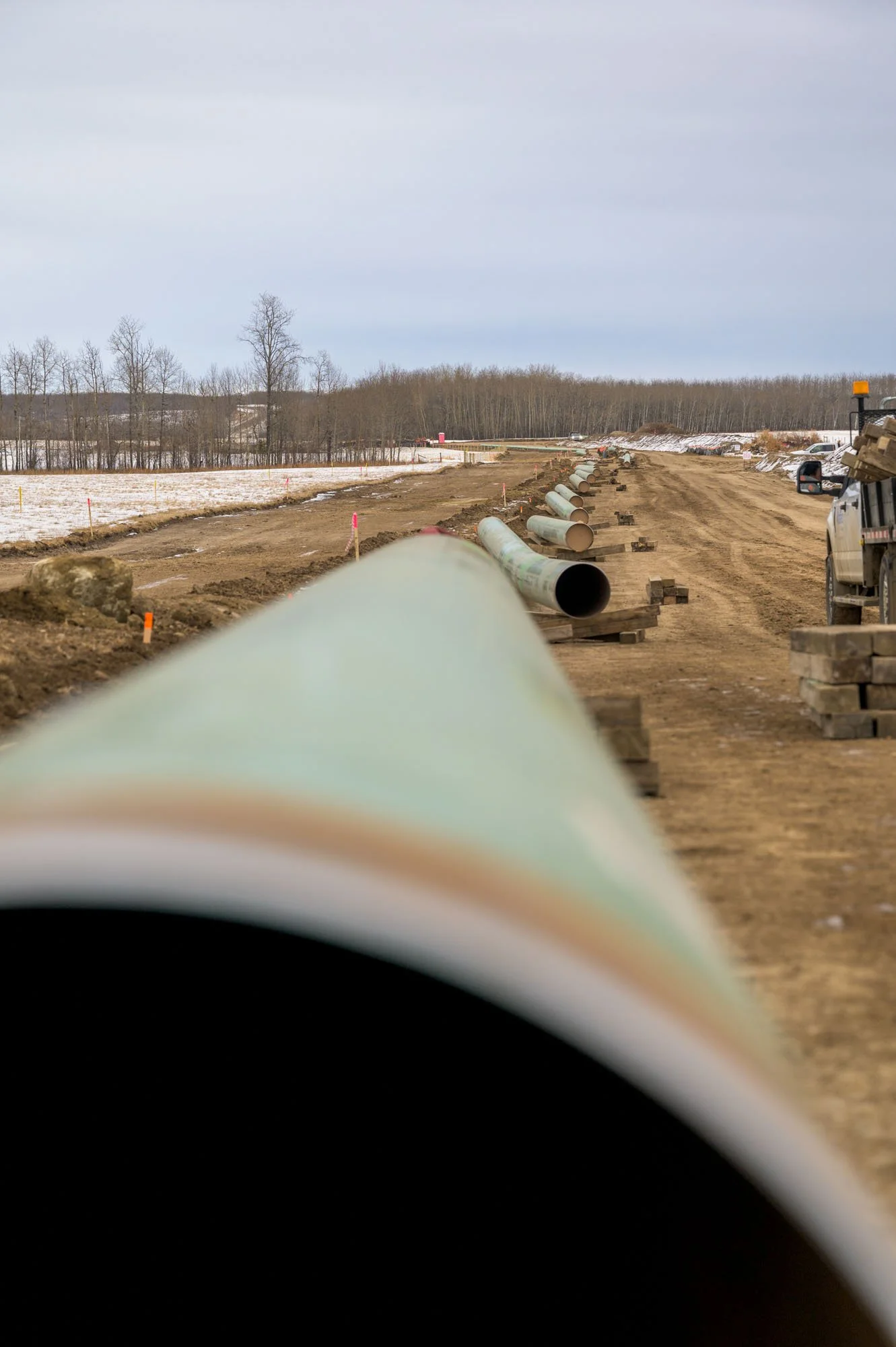 Construction site with pipes laid out along a dirt road, snow on the ground, and a truck parked on the right.