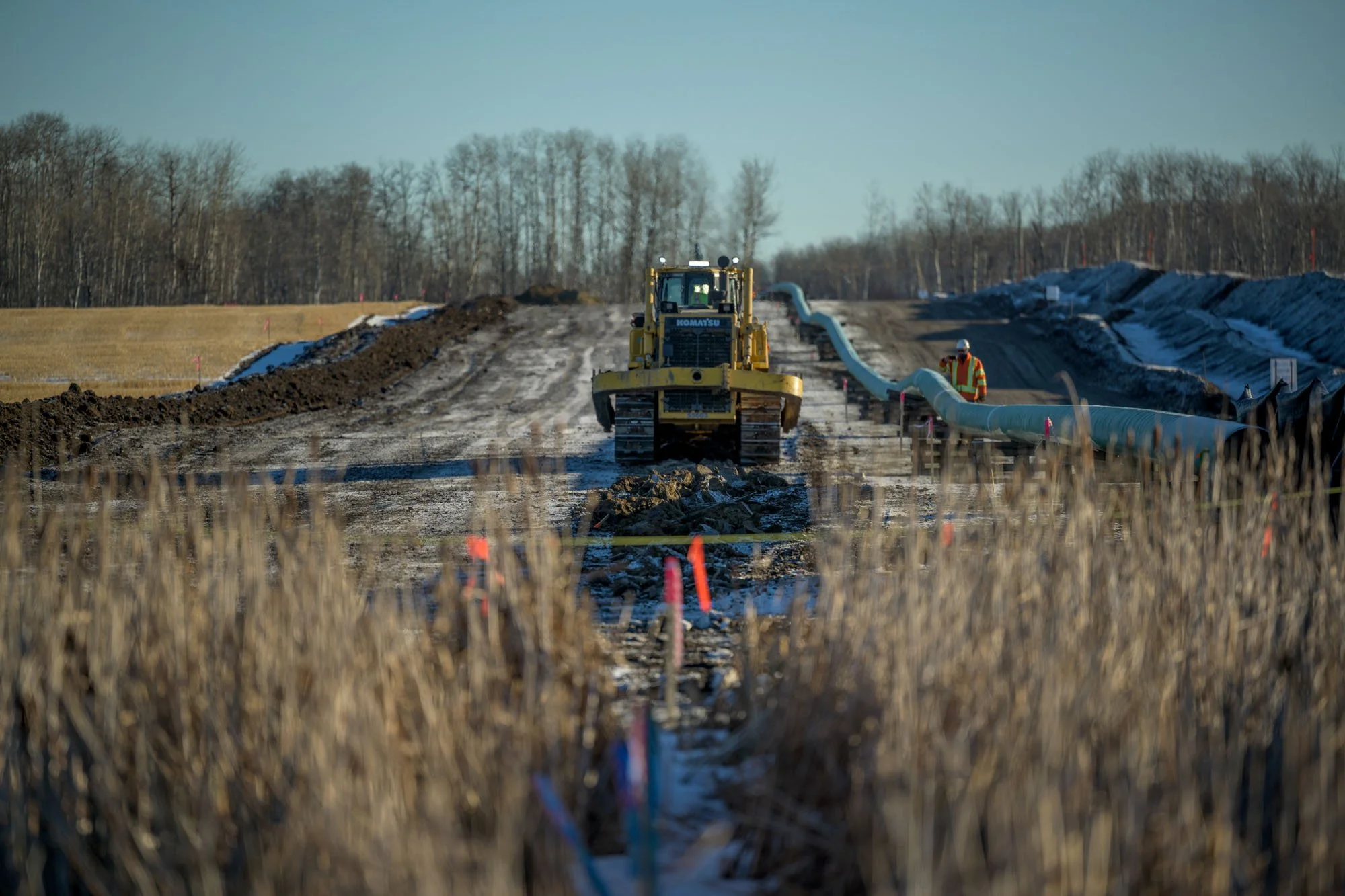Construction workers and machinery on a pipeline installation site in a rural area during winter, with trees in the background.