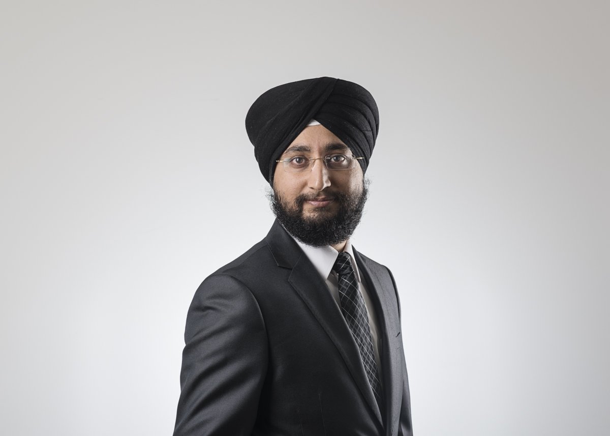 A man wearing glasses, a black turban, a black suit, white shirt, and striped tie posing against a light gray background.
