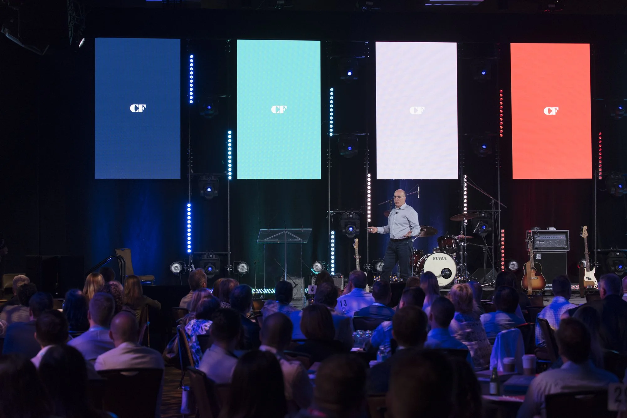 A man on stage giving a presentation to a large audience at a conference with four large vertical screens behind him displaying different colors with the initials "CF".