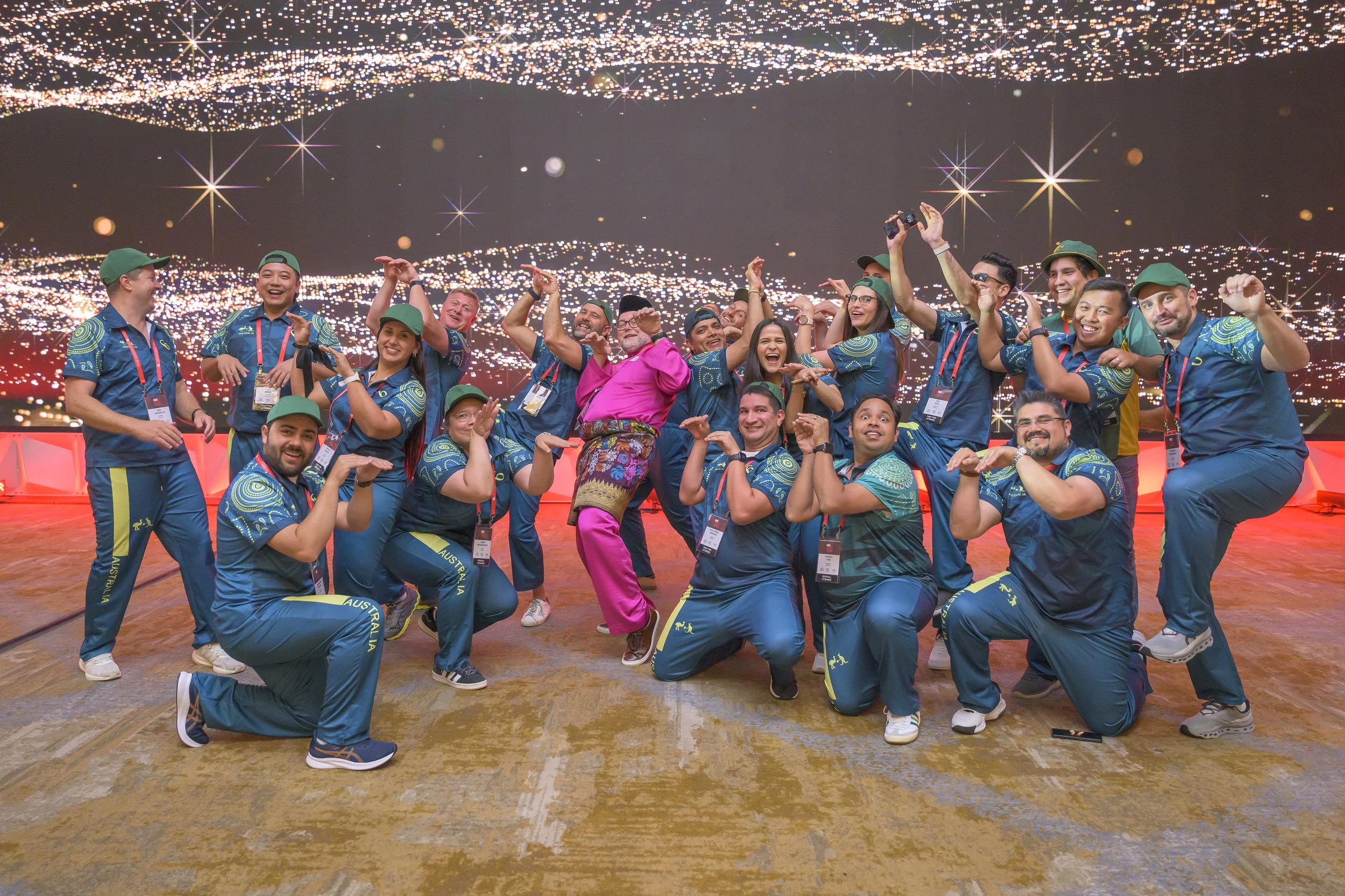 A diverse group of people wearing blue Australian team uniforms, posing together and smiling at an event, with a city-lit backdrop and many stars in the sky.