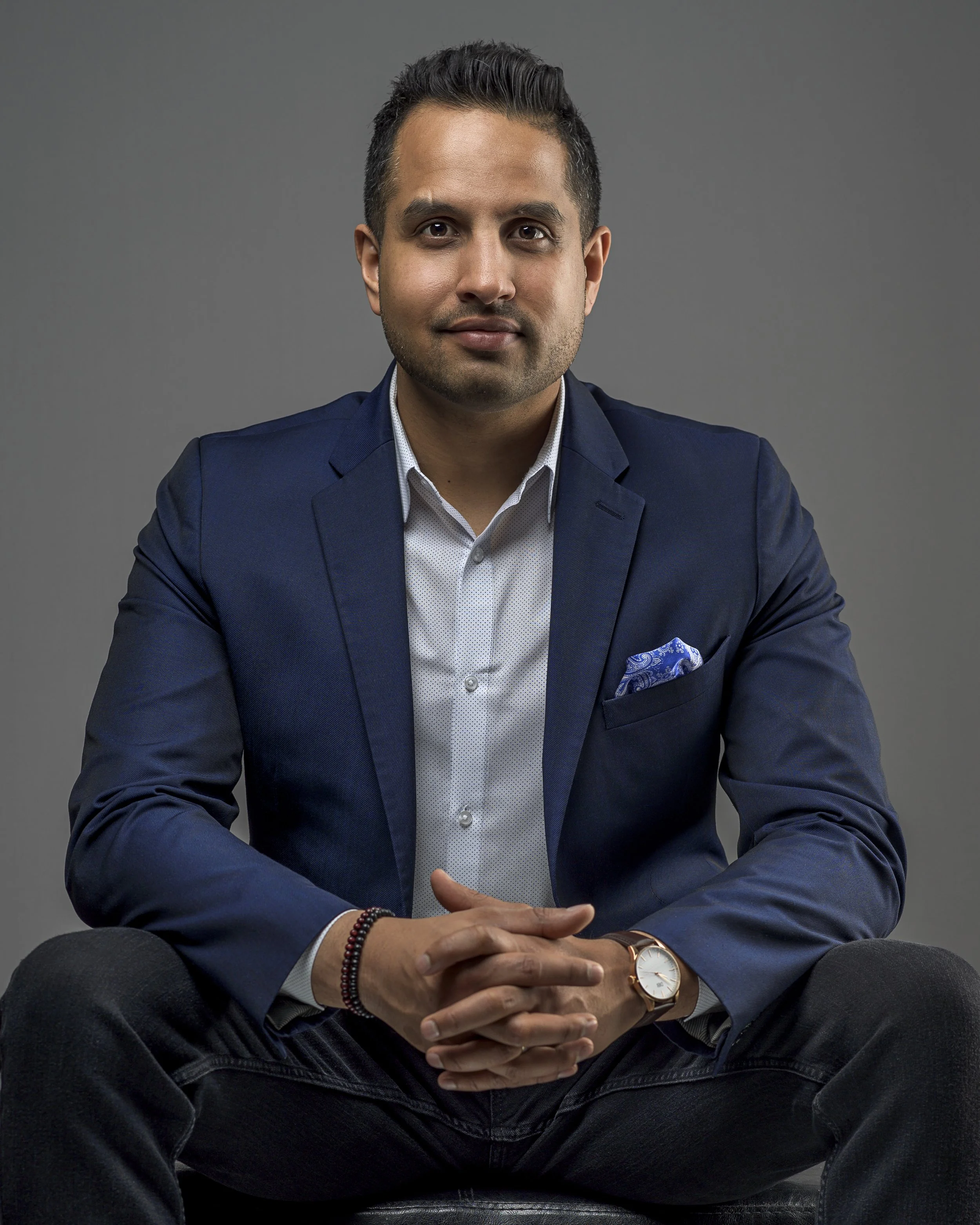 Portrait of a man in a navy blue suit jacket, white shirt, and casual pants, seated with hands clasped, against a plain gray background.