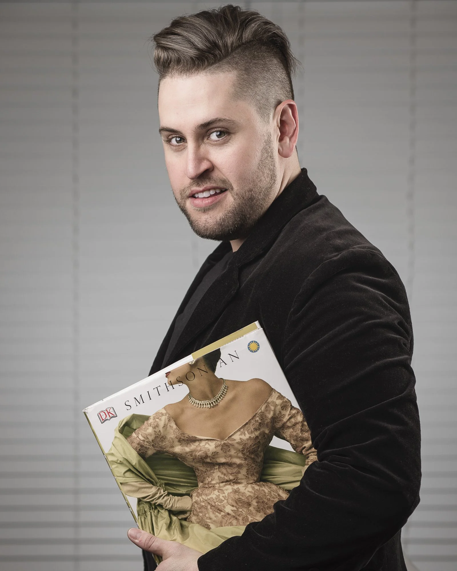 A man with styled hair and a beard holding a book, standing in front of a plain background.