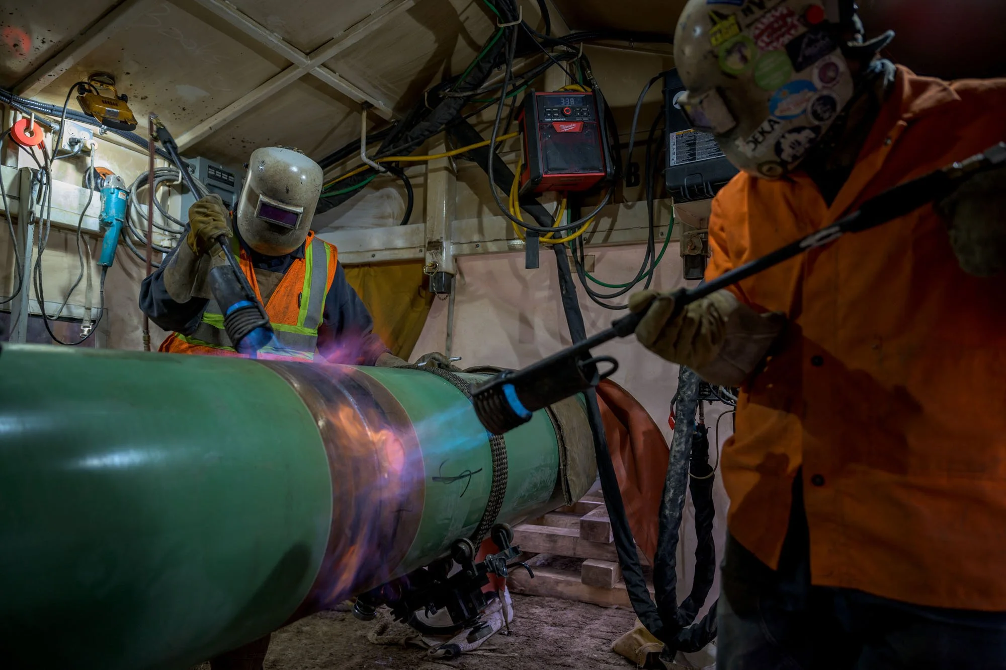 Two workers wearing protective gear and orange vests are welding a large green pipe inside a construction or industrial site, with welding sparks visible.