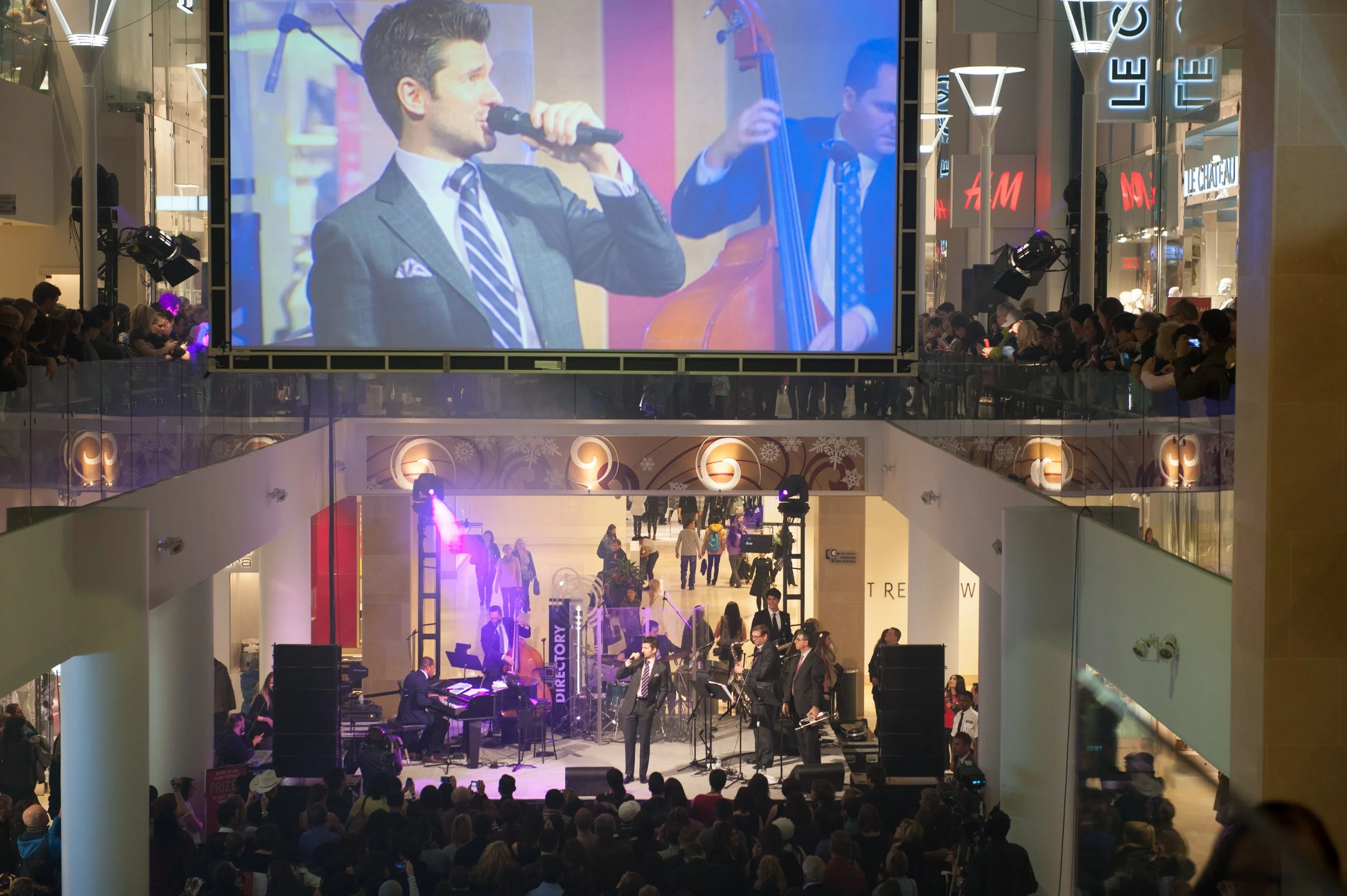 A live jazz band performing on a stage inside a shopping mall with an audience, a large screen above showing the band, and shoppers on the upper level watching.