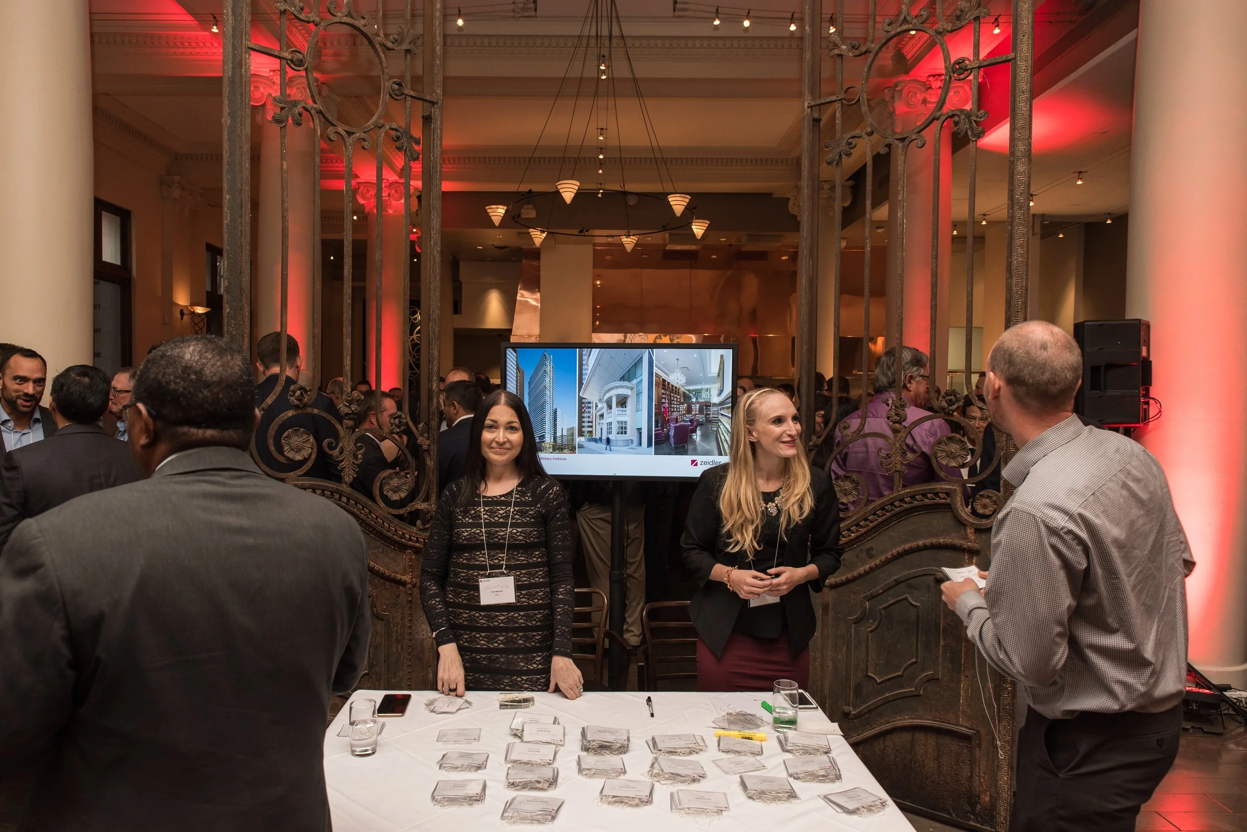 People at a professional event or conference, with a table displaying name tags and a large screen showing building images in the background.