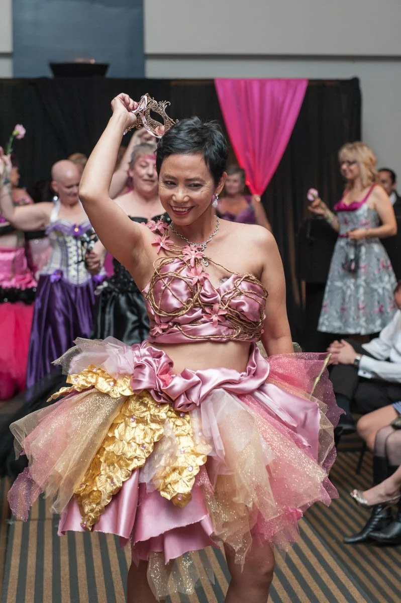 A woman in a pink and gold tutu dress holds a crown above her head at a party, smiling, with other women in colorful dresses in the background.