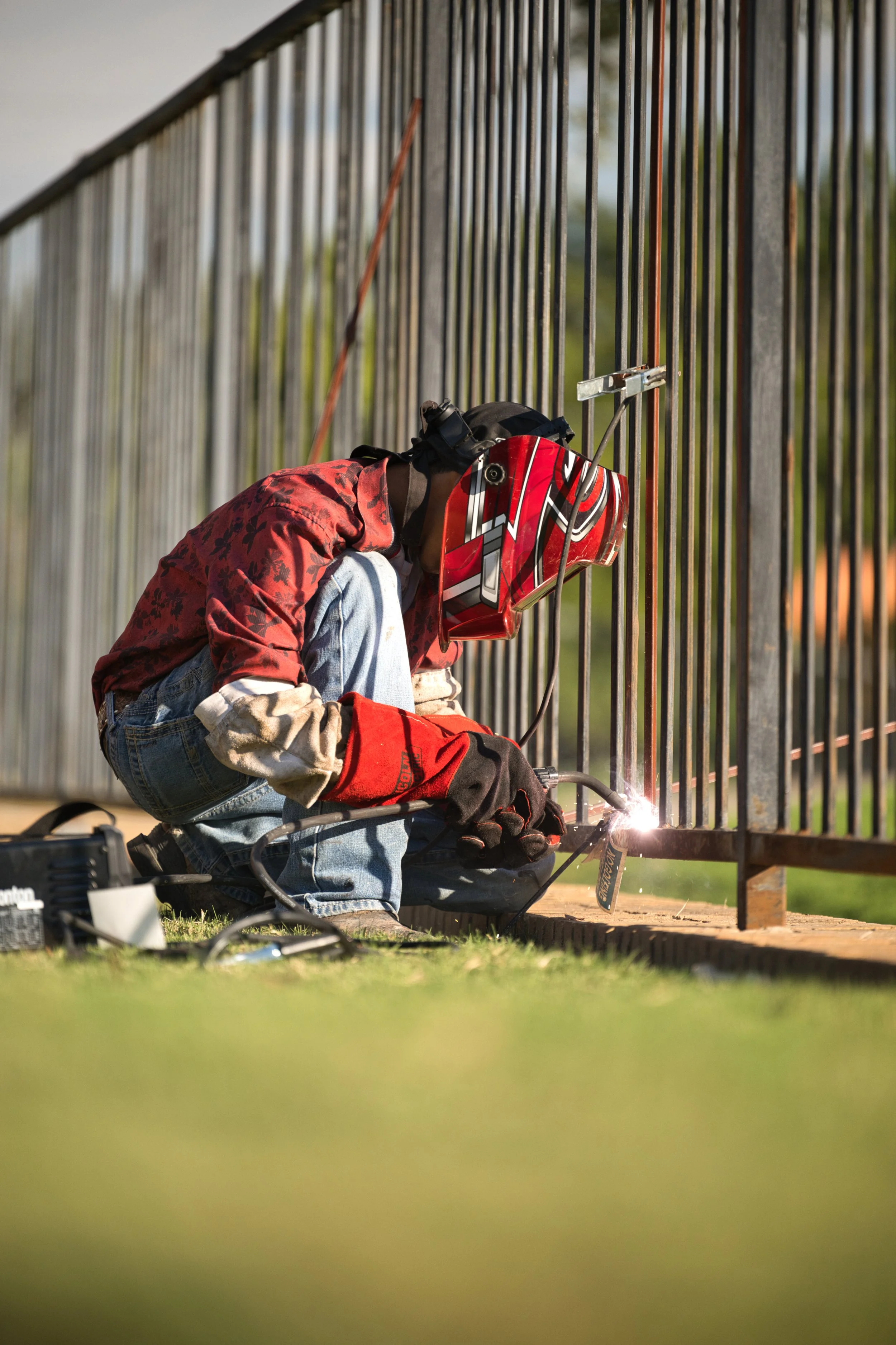 A person welding metal bars of a fence while kneeling on the ground, wearing a red and black helmet, red gloves, a red jacket, and blue jeans.