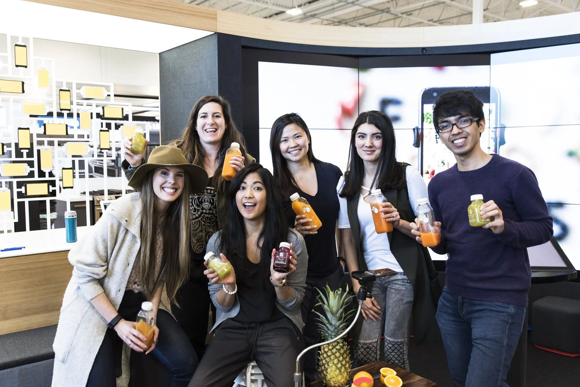 Group of diverse young adults smiling and holding bottles of juice at a social event in a modern space with digital screens in the background.