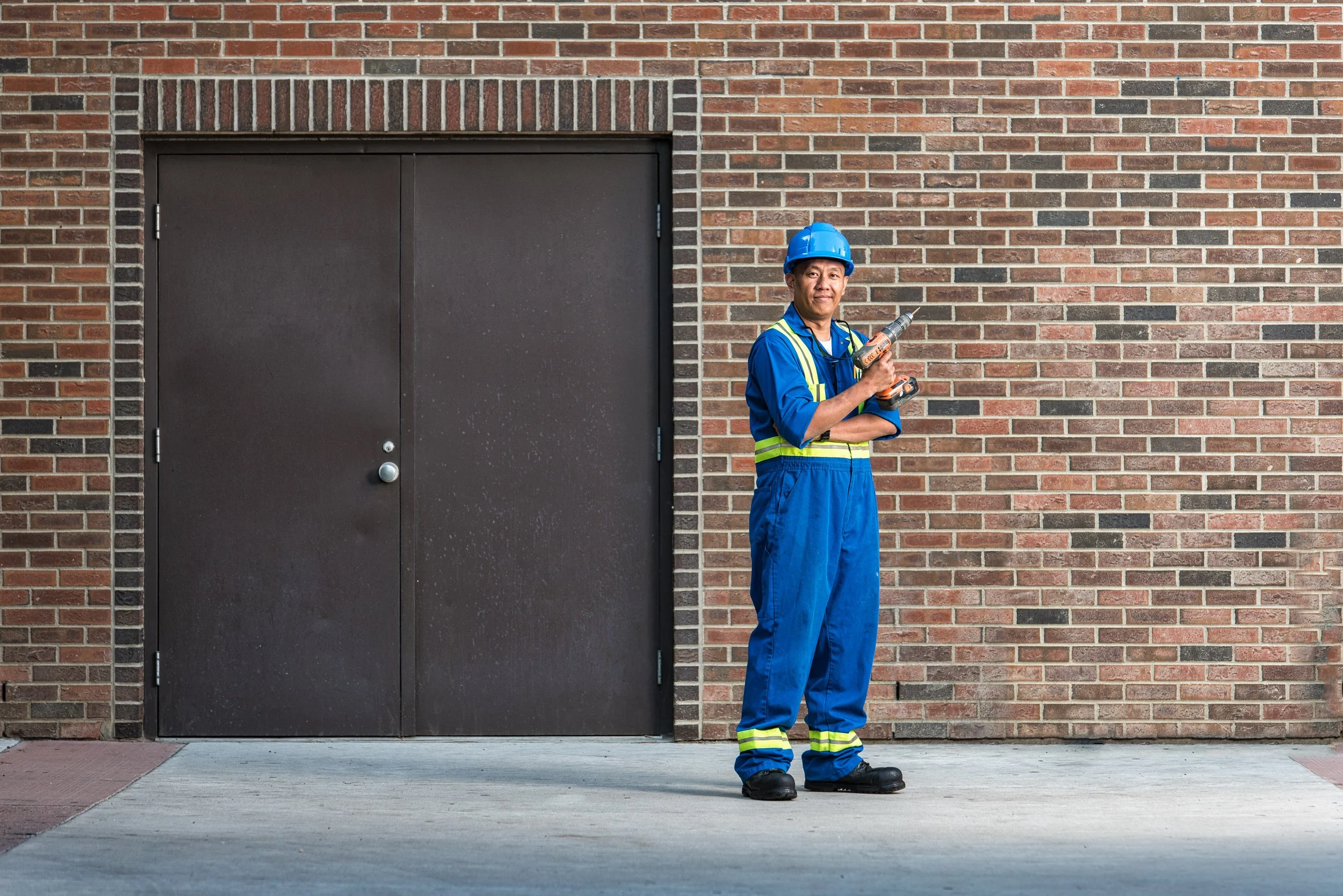 A construction worker in blue overalls and a helmet standing outside near a brown brick wall and a dark door, holding a cordless drill.