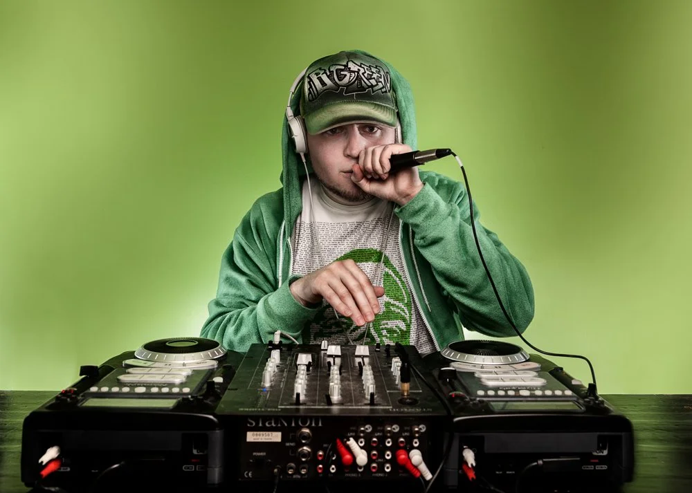A young man wearing a green hoodie, cap, and headphones is holding a microphone and DJing with a mixer and turntables in front of a green background.