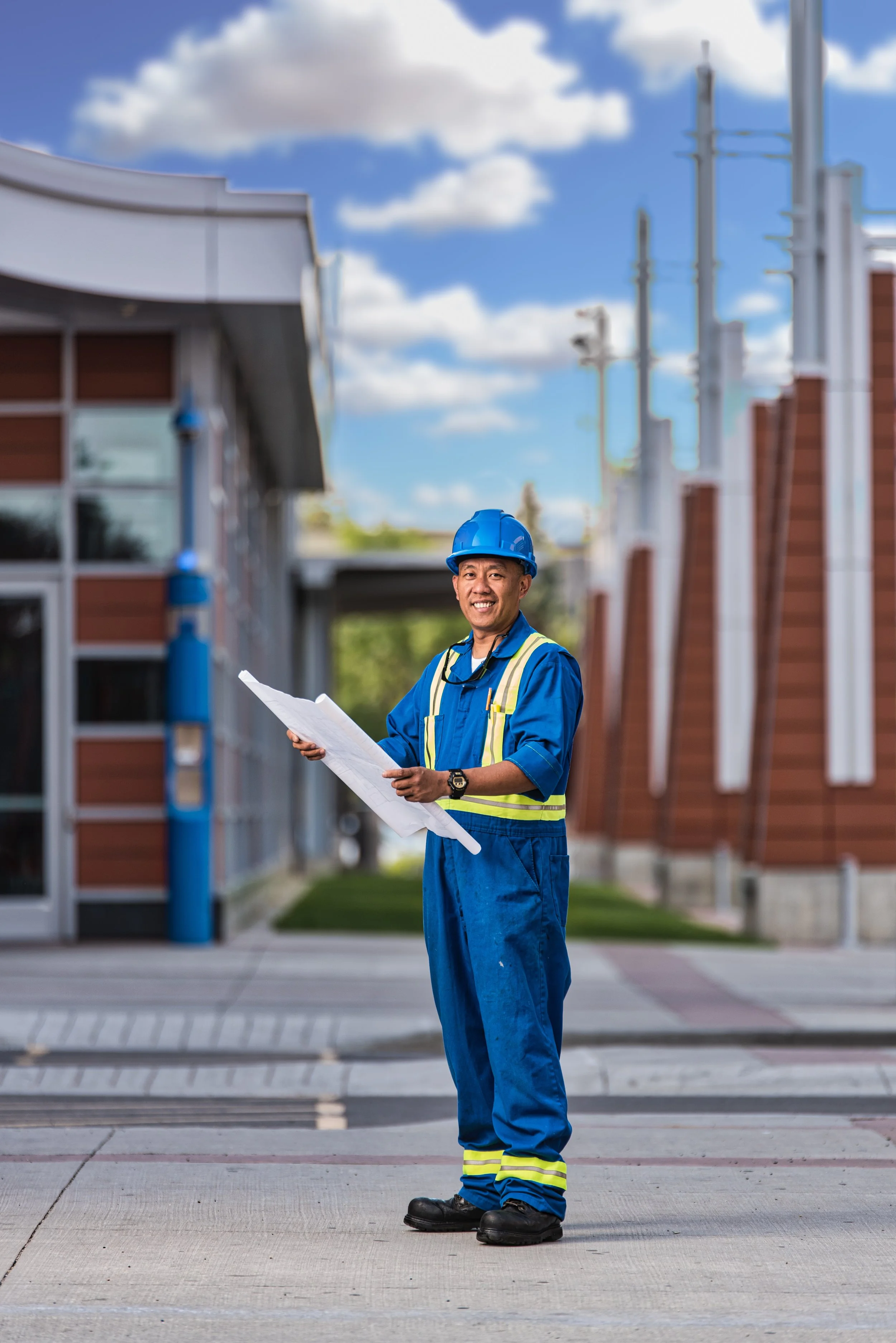 A construction worker wearing a blue safety helmet and blue uniform with yellow reflective stripes, holding blueprints and smiling outdoors on a sunny day with a modern building in the background.