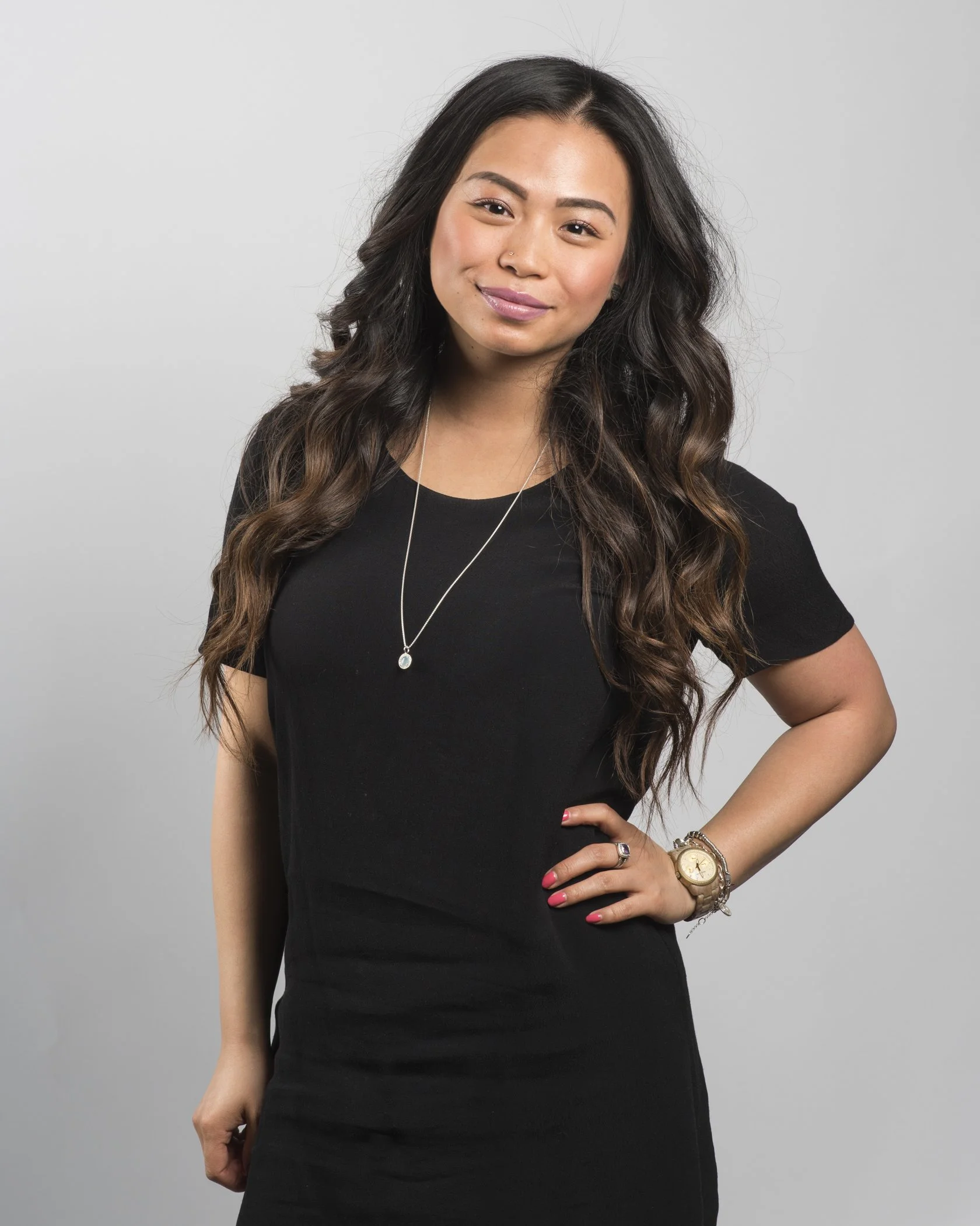 A woman with long, wavy dark hair, wearing a black dress, a necklace, a watch, and a ring, posing with her hand on her hip against a plain gray background.