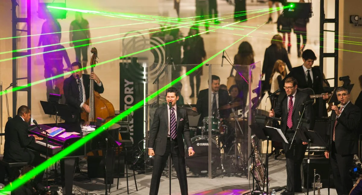 Jazz band performing on stage with green laser lights and a crowd in the background.