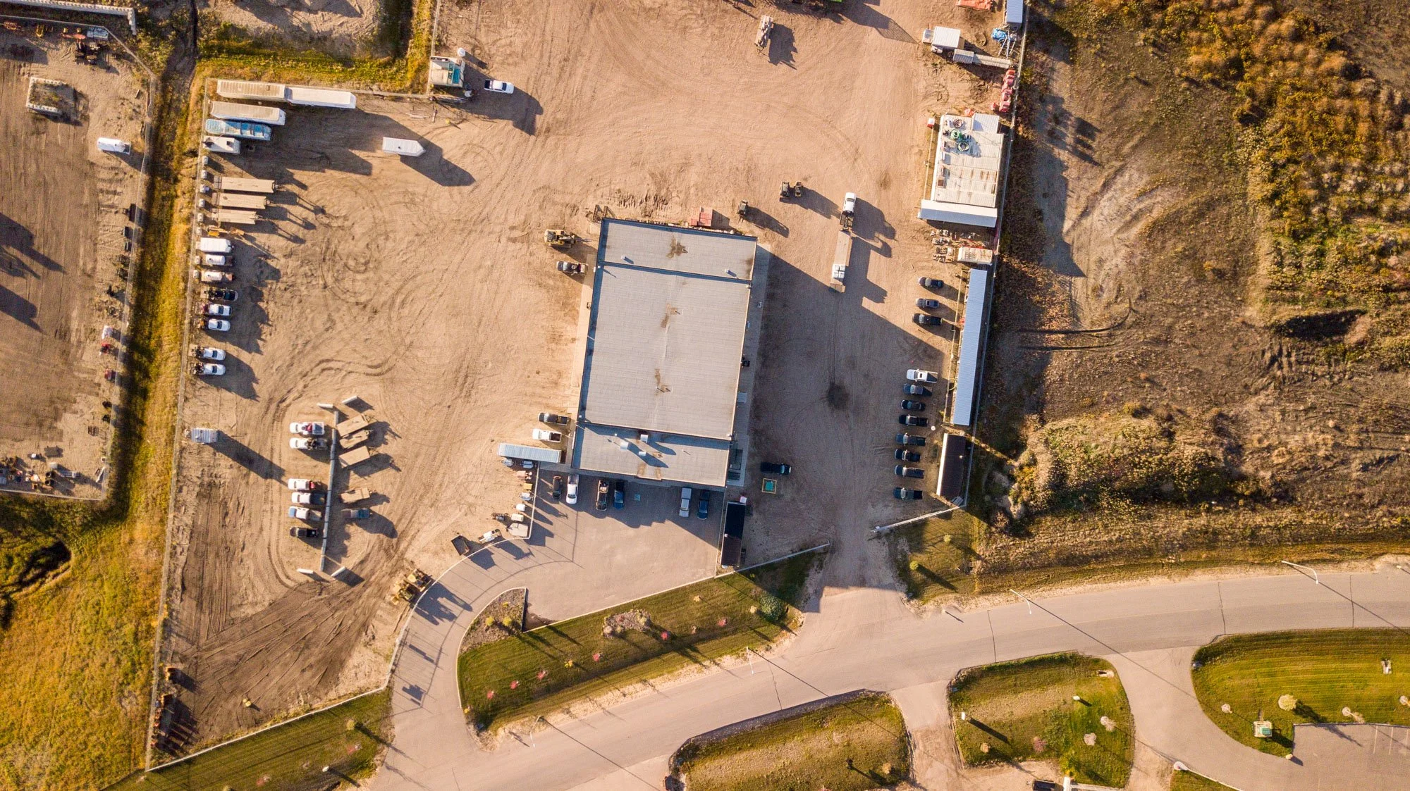 An aerial view of a mostly empty parking lot and a large building, with some cars parked near the building, dirt areas, and surrounding greenery.