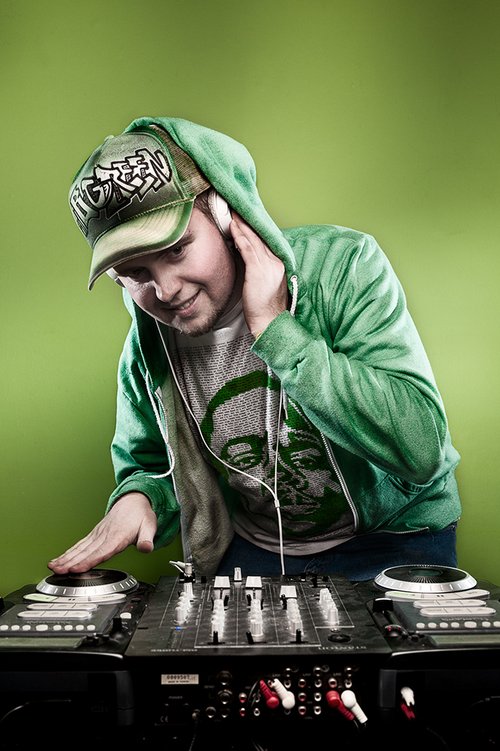 A young man in a green hoodie, cap, and headphones, DJing with a mixer and turntables against a green background.