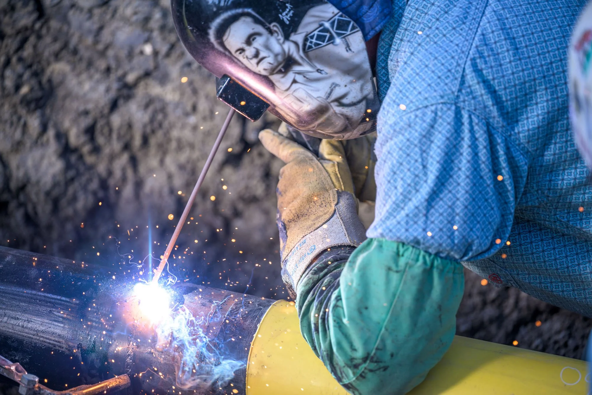 A worker welding a yellow metal pipe using a torch, with sparks and bright light.
