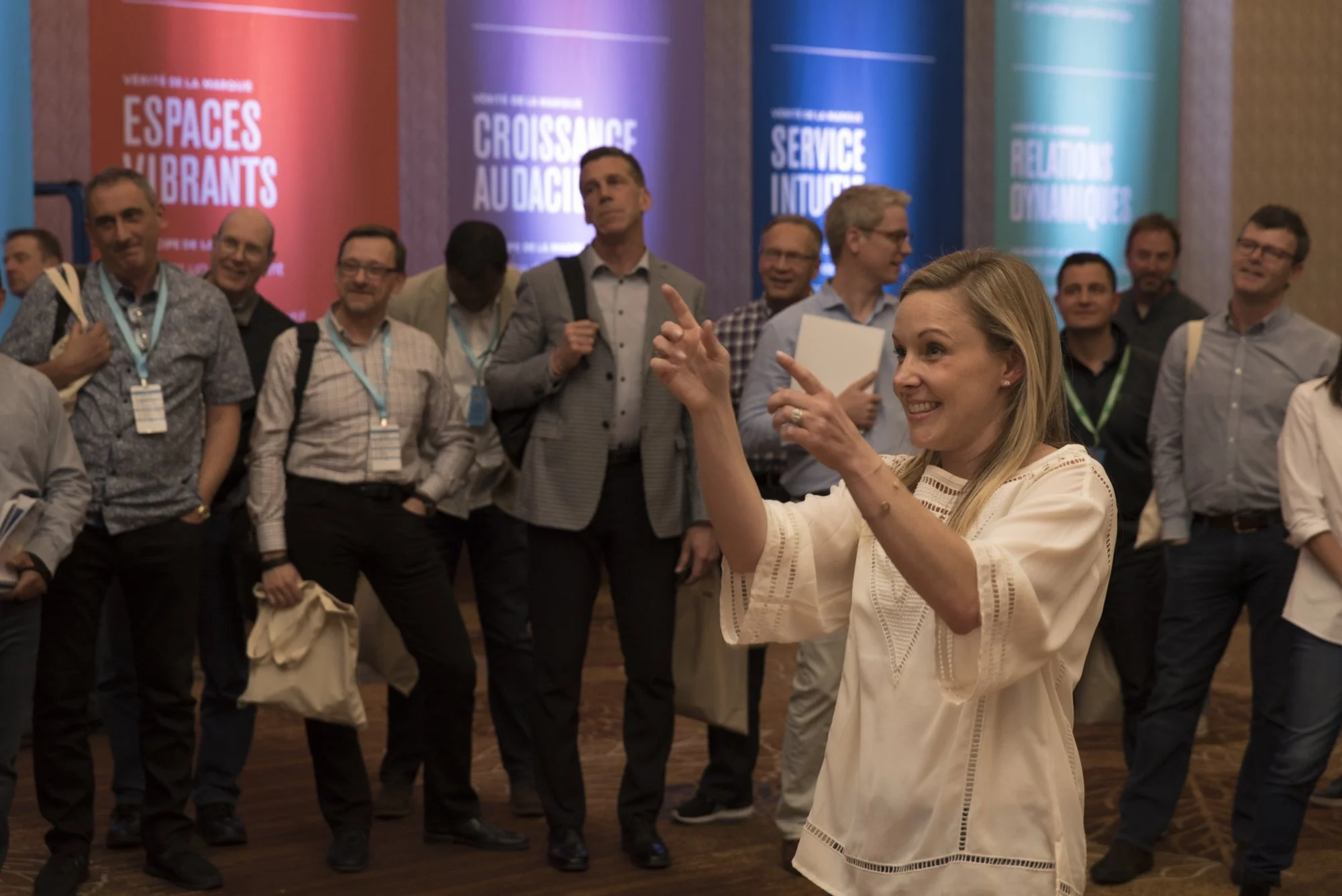 A woman is speaking and gesturing in front of a group of men at a conference, with colorful banners in French in the background.