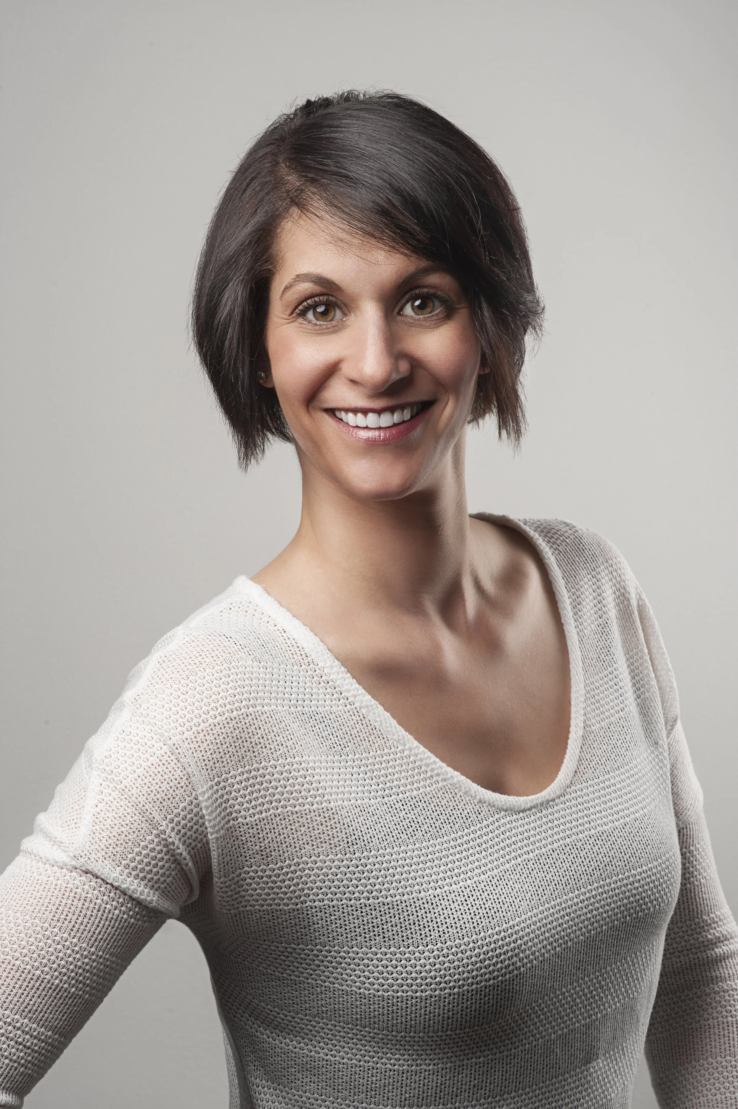 A woman smiling with short brown hair wearing a white knitted top against a plain gray background.