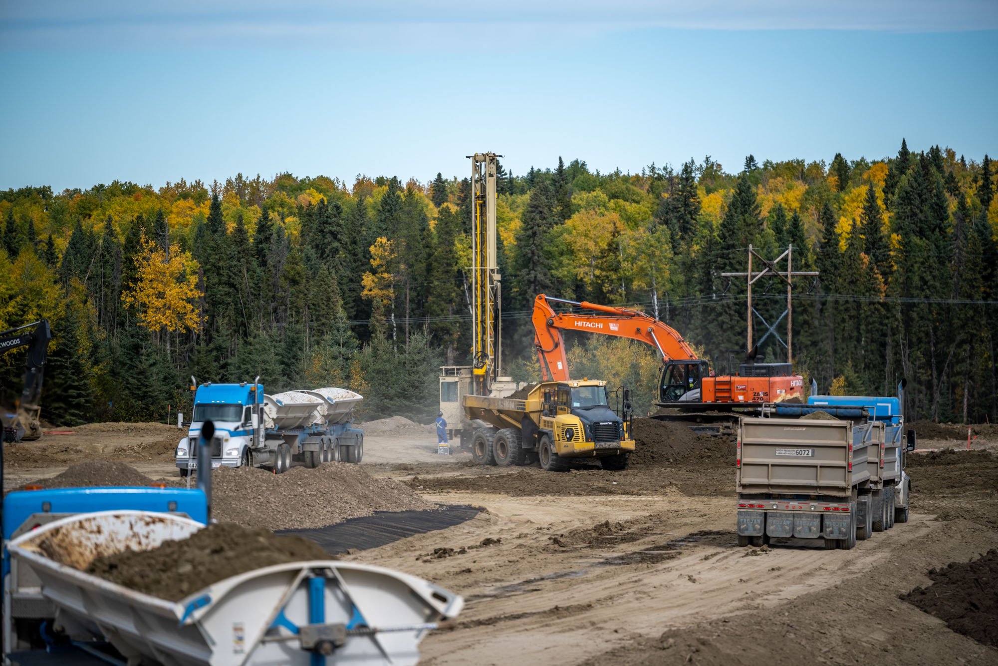 Construction site with heavy machinery including excavators, trucks, and drilling equipment, with a forest in the background and a dirt road in the foreground.