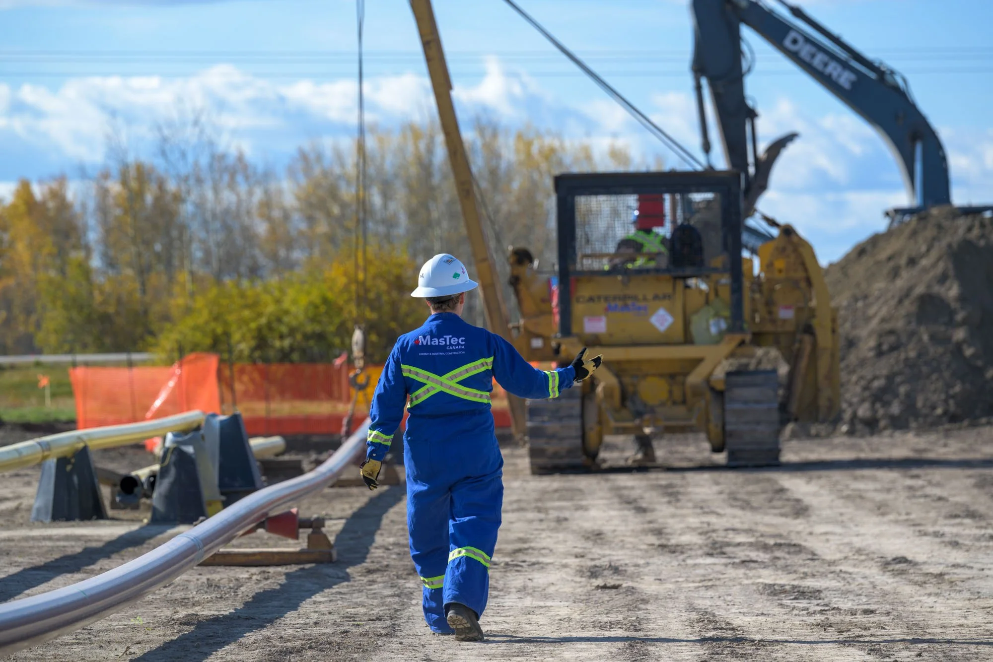 Construction worker in blue safety gear and white helmet walking at a construction site with heavy machinery and pipelines.