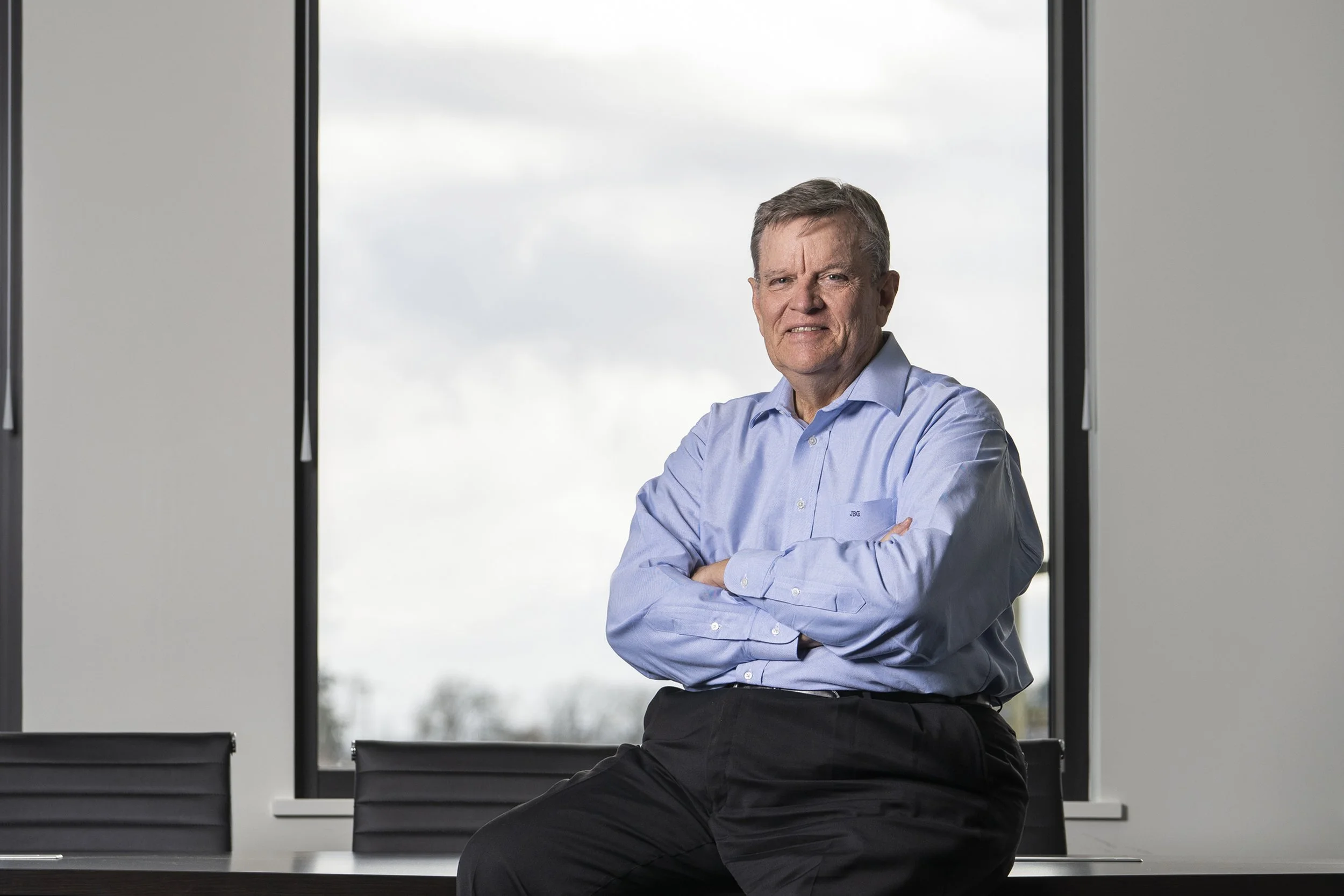 A middle-aged man in a light blue dress shirt and dark pants sitting on the edge of a conference table with arms crossed, inside a modern office with large window and cloudy sky outside.