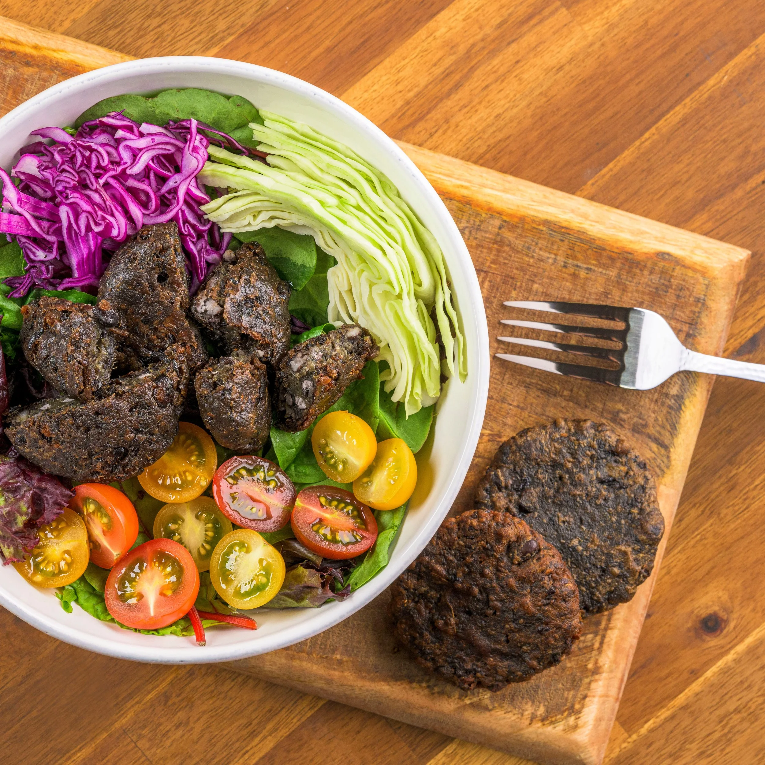 A bowl of salad with cherry tomatoes, shredded purple cabbage, sliced cucumber, and dark meat chunks on a wooden cutting board with two cooked veggie patties and a fork.